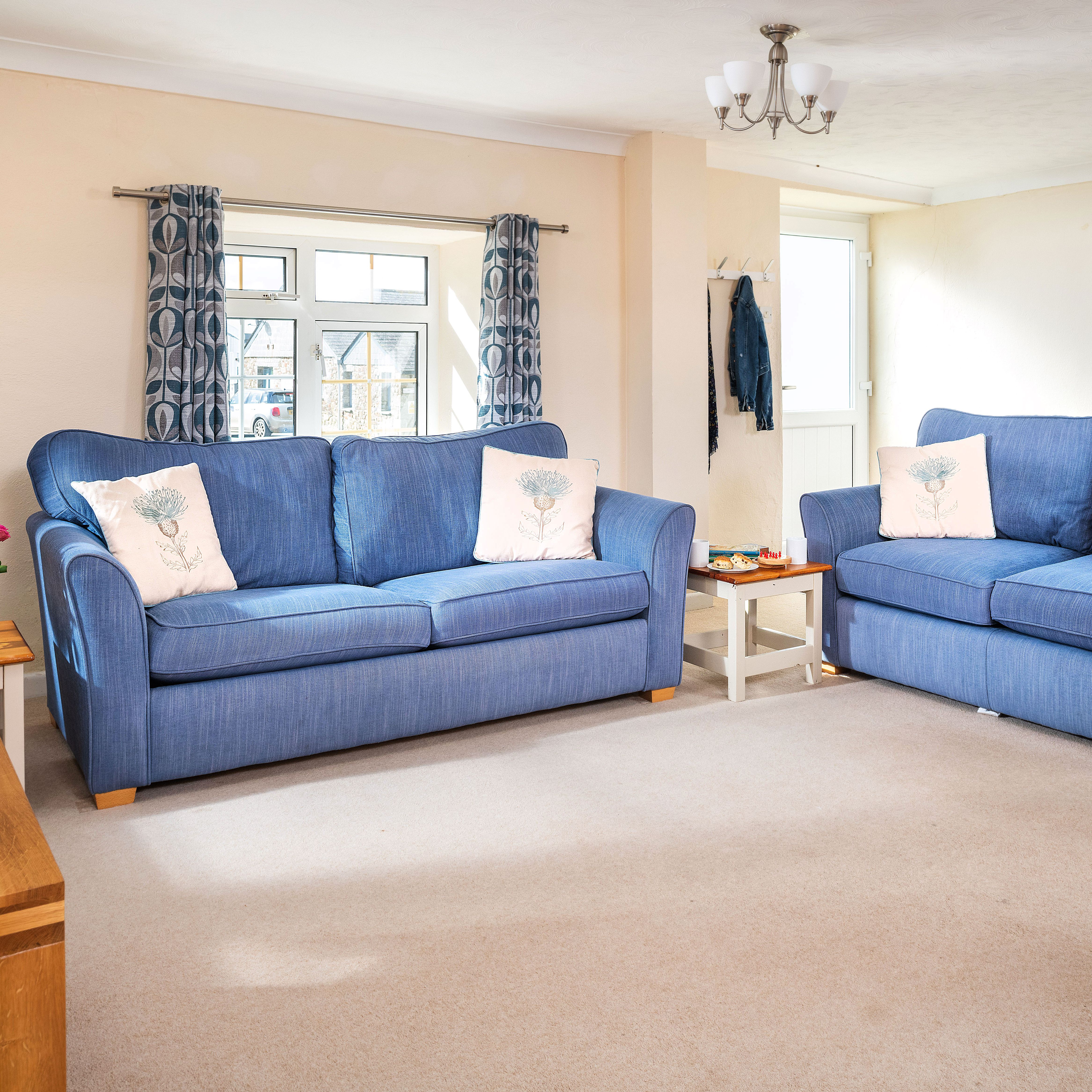 A bright and cozy living room with two blue sofas, decorative pillows, a clock on the wall, patterned curtains, a side table with a vase of flowers, and wall art featuring flying birds.
