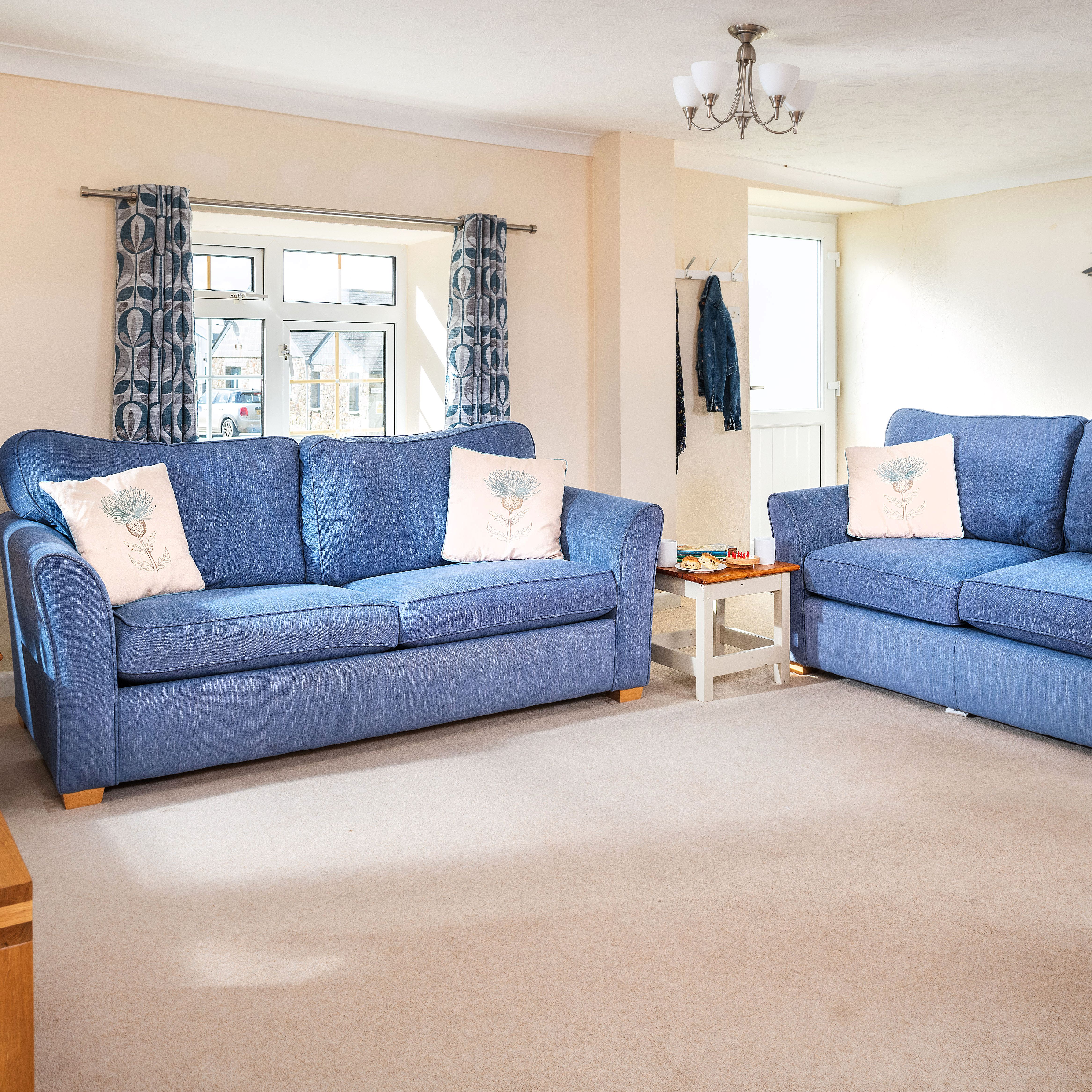A bright and cozy living room with two blue sofas, decorative pillows, a clock on the wall, patterned curtains, a side table with a vase of flowers, and wall art featuring flying birds.
