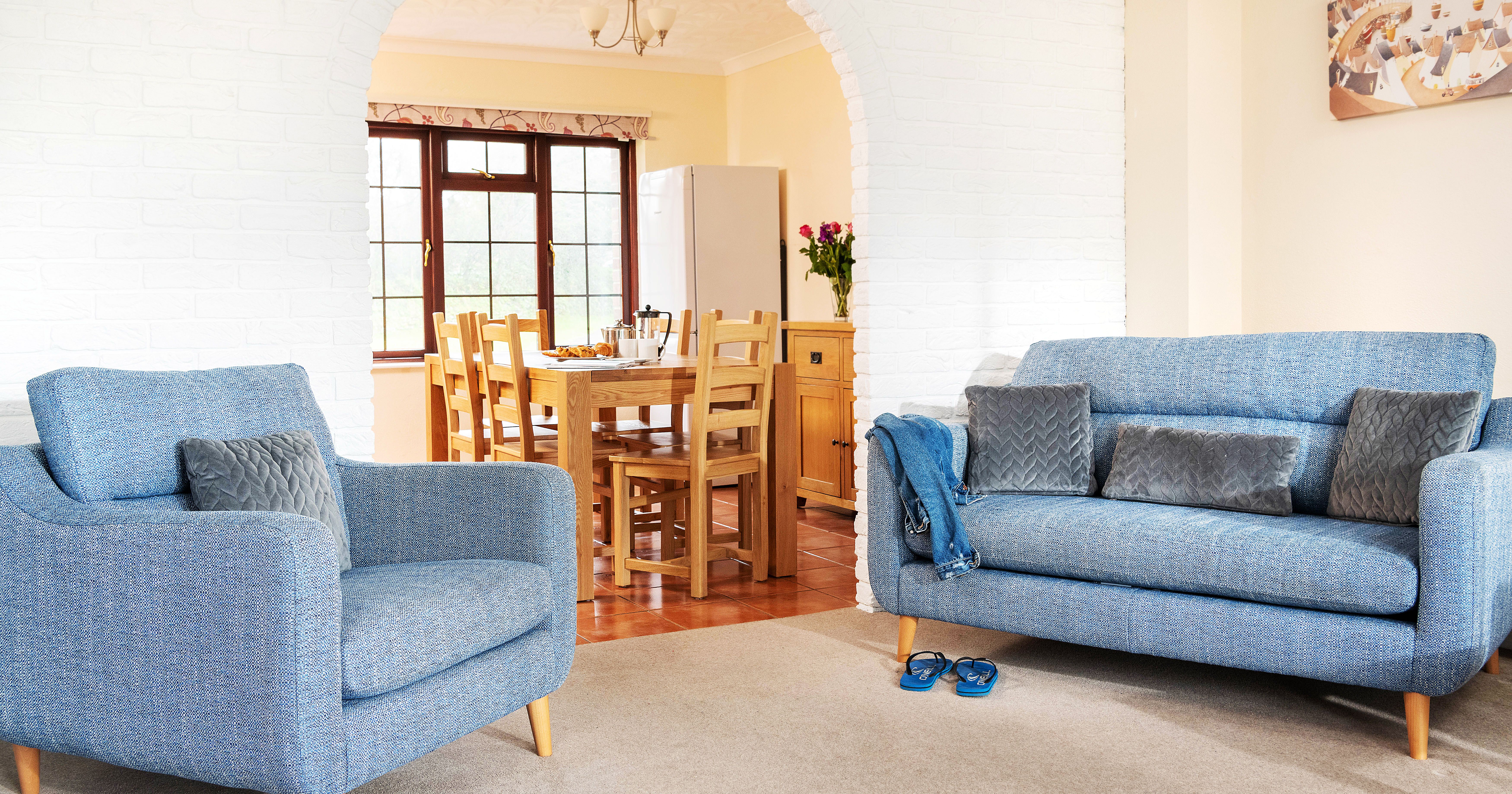 Bright living room with blue upholstered armchair and sofa, gray cushions, blue slippers, and a dining area with a wooden table and chairs visible through an arched doorway.