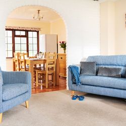 Bright living room with blue upholstered armchair and sofa, gray cushions, blue slippers, and a dining area with a wooden table and chairs visible through an arched doorway.