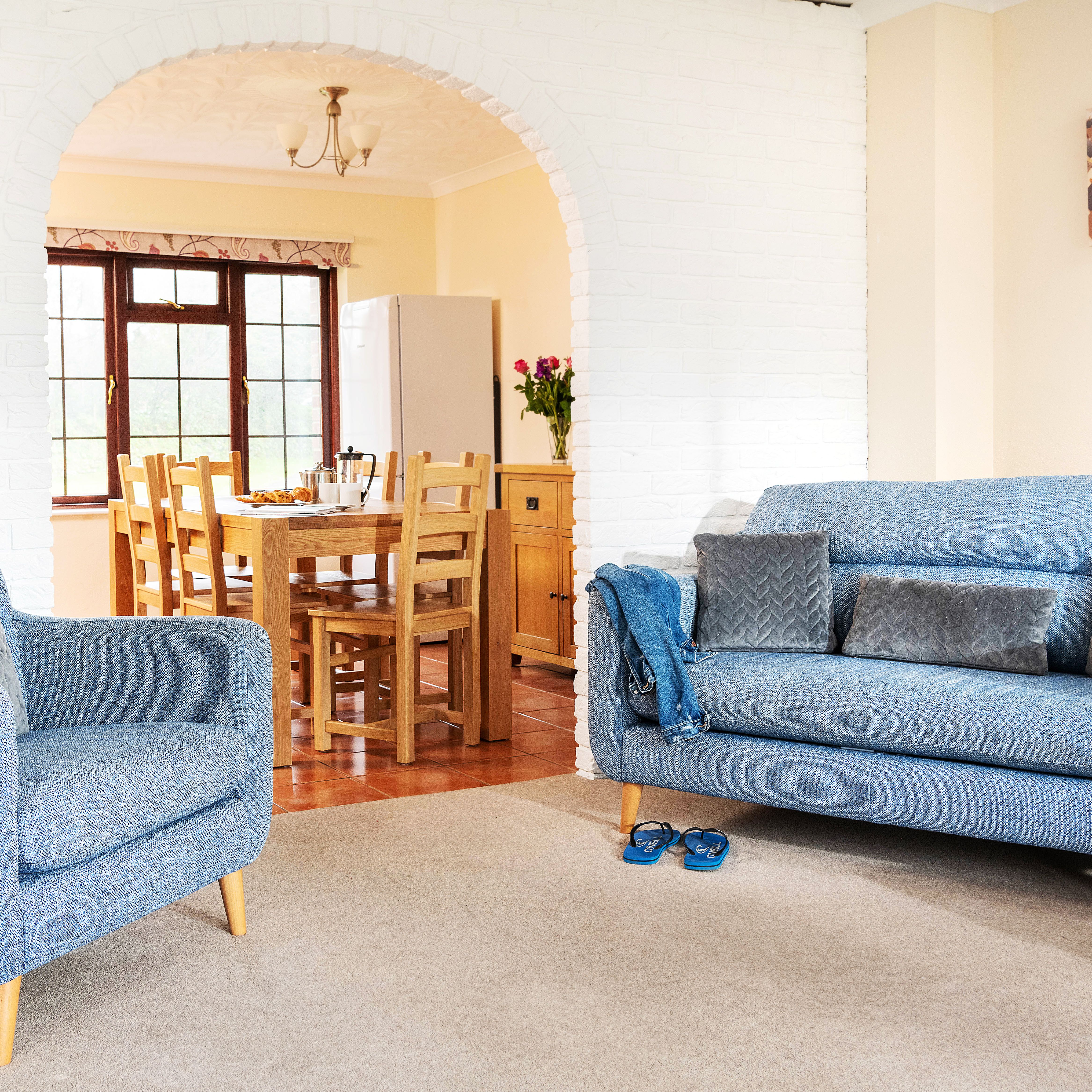 Bright living room with blue upholstered armchair and sofa, gray cushions, blue slippers, and a dining area with a wooden table and chairs visible through an arched doorway.