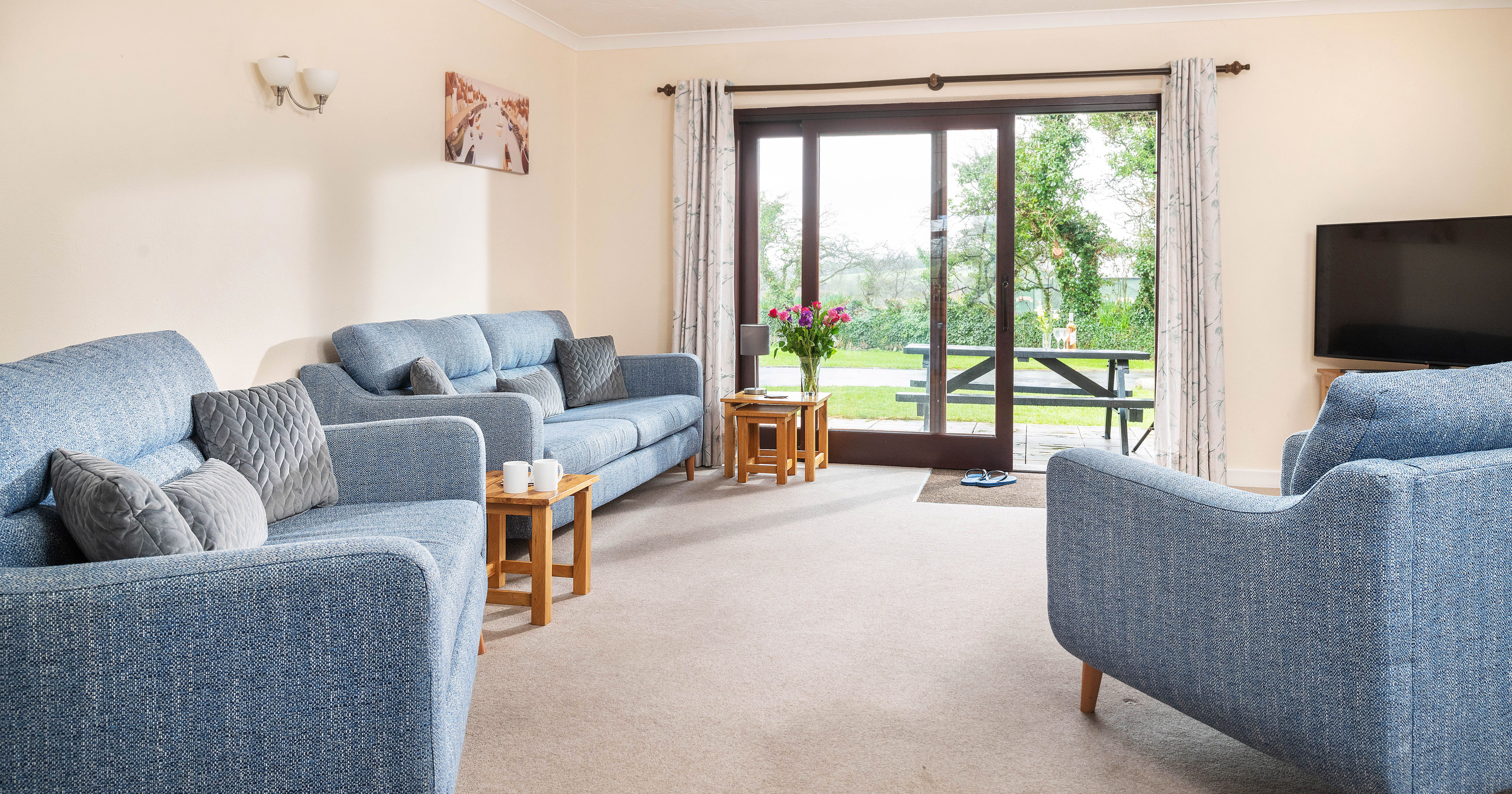 Bright living room with blue sofas, wooden coffee tables, large TV, and sliding glass doors opening to a garden view.