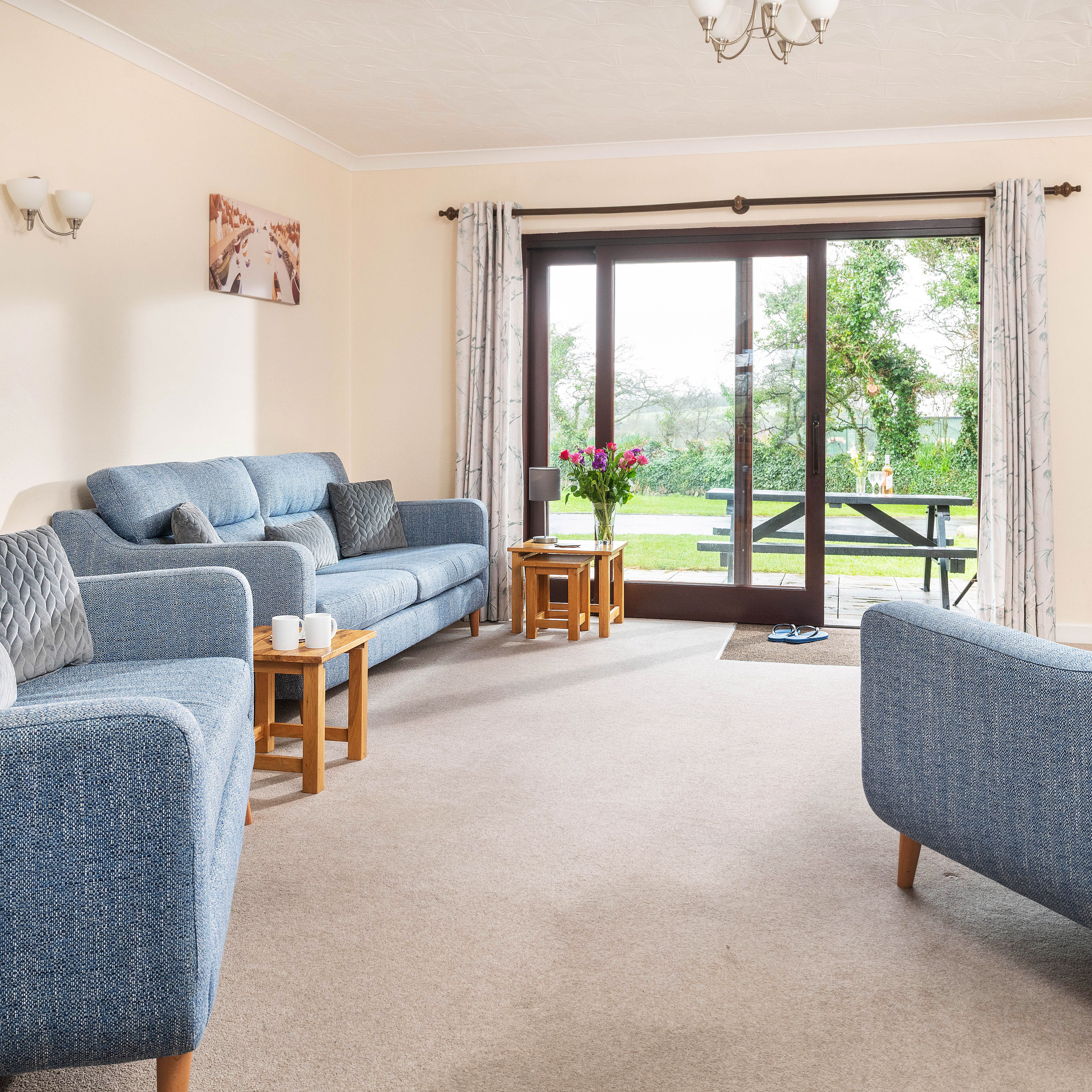 Bright living room with blue sofas, wooden coffee tables, large TV, and sliding glass doors opening to a garden view.