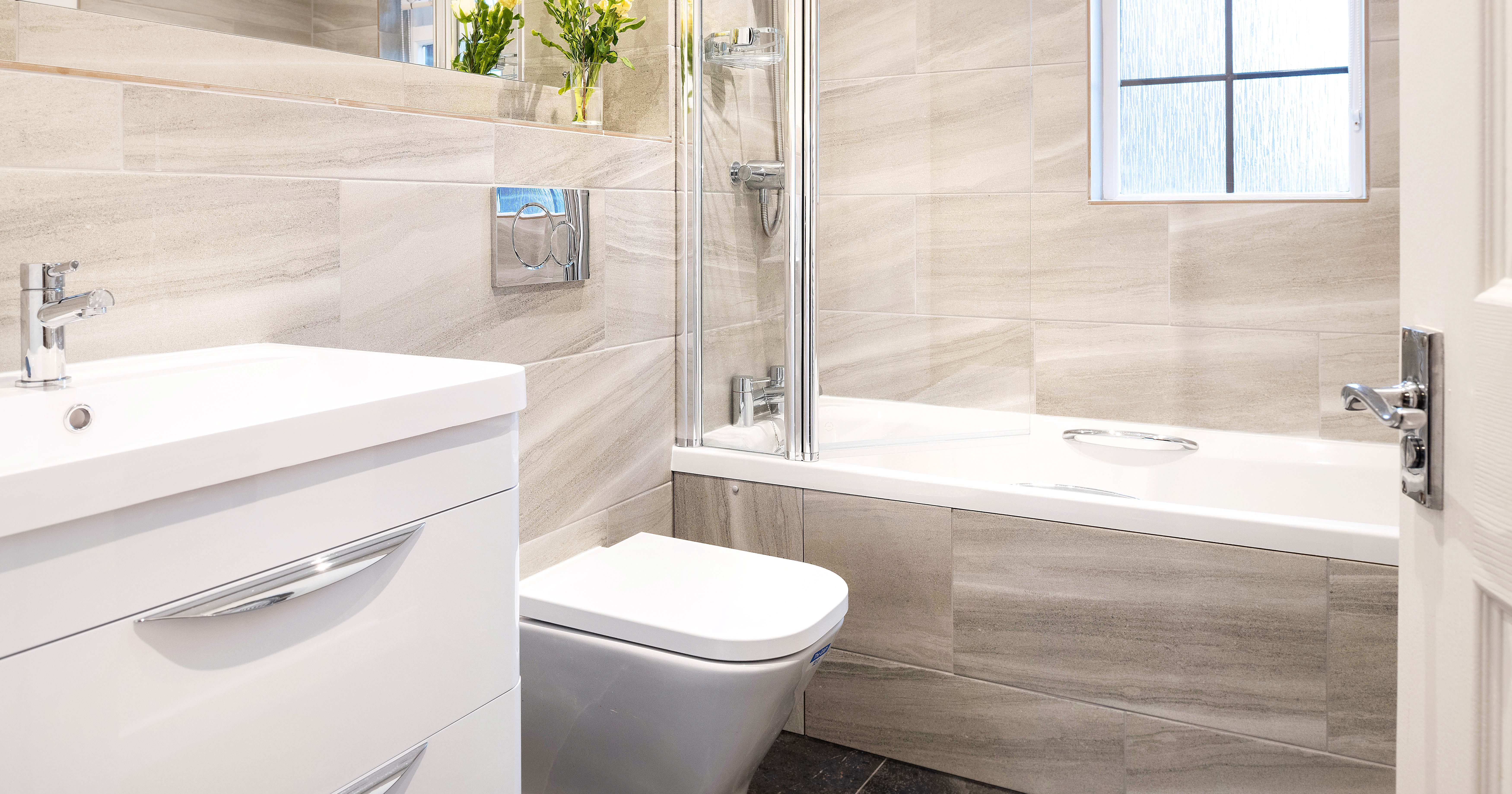 Modern bathroom with beige tiles, a white vanity sink, a toilet, a combined bathtub and shower, and a window. There are yellow flowers placed near the mirror.