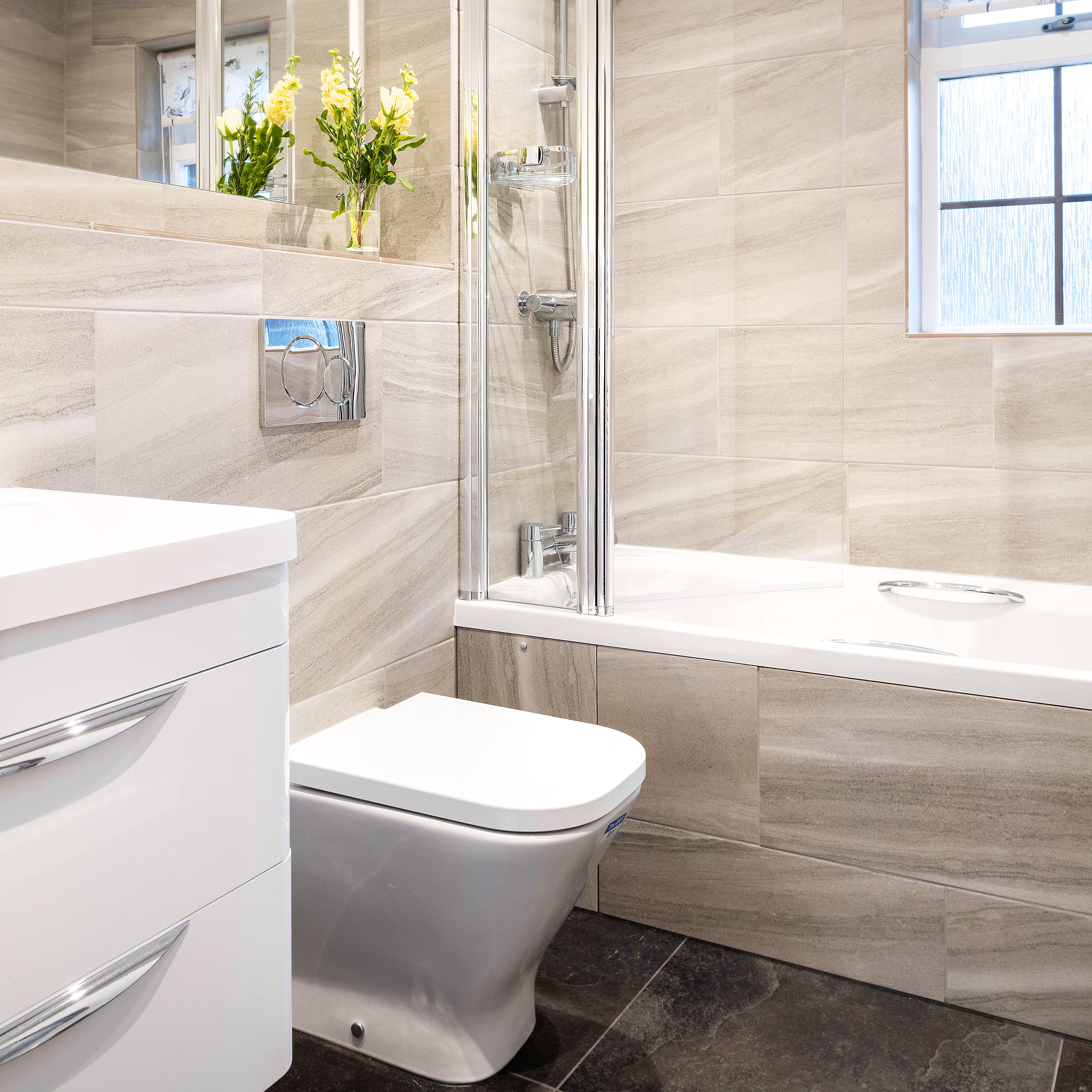 Modern bathroom with beige tiles, a white vanity sink, a toilet, a combined bathtub and shower, and a window. There are yellow flowers placed near the mirror.