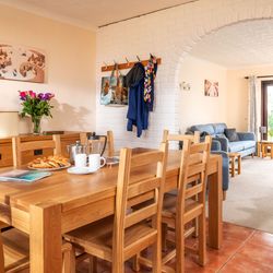 Bright open-plan dining and living room with wooden table and chairs, blue sofa, flowers on a sideboard, coat rack, and large glass doors leading to a garden.