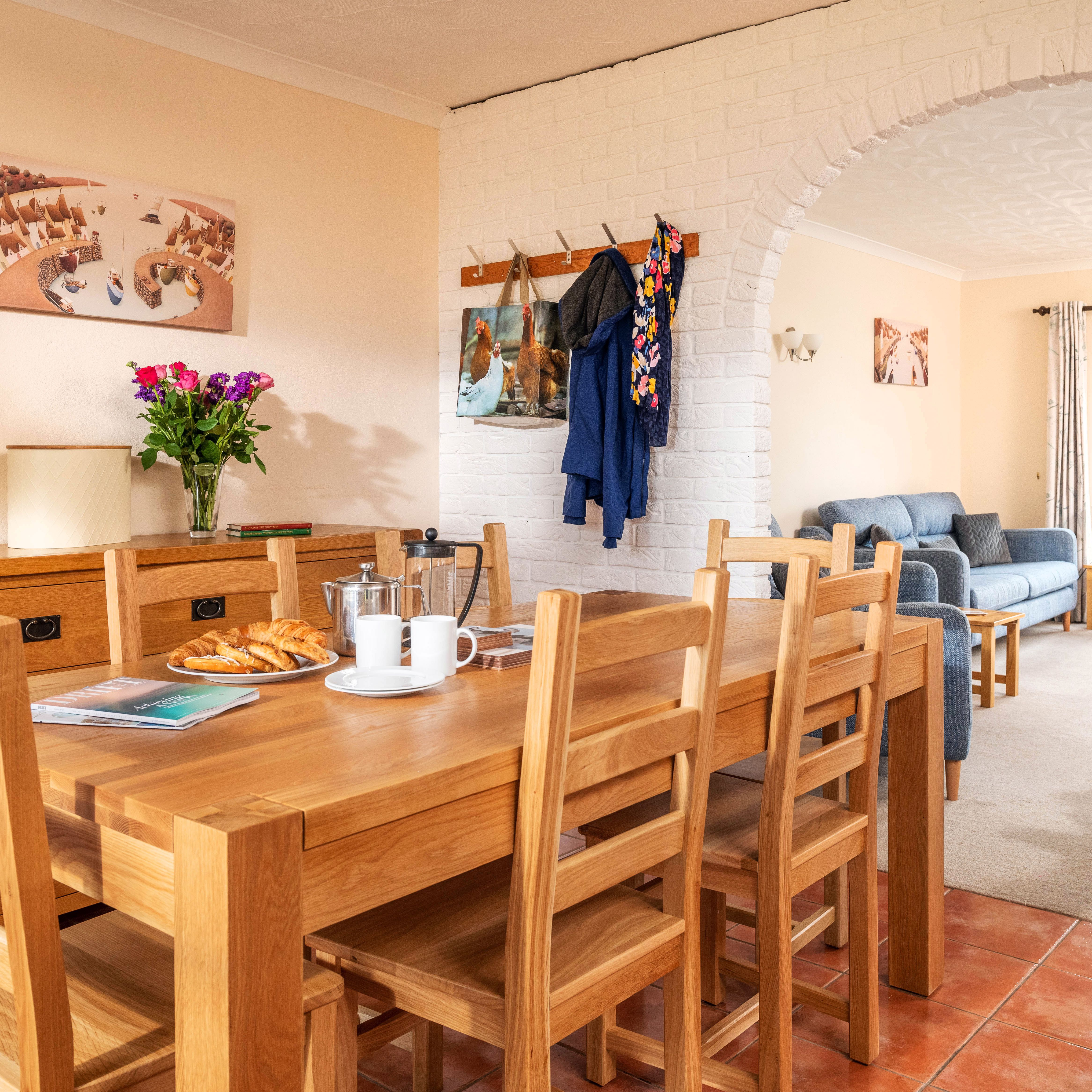 Bright open-plan dining and living room with wooden table and chairs, blue sofa, flowers on a sideboard, coat rack, and large glass doors leading to a garden.