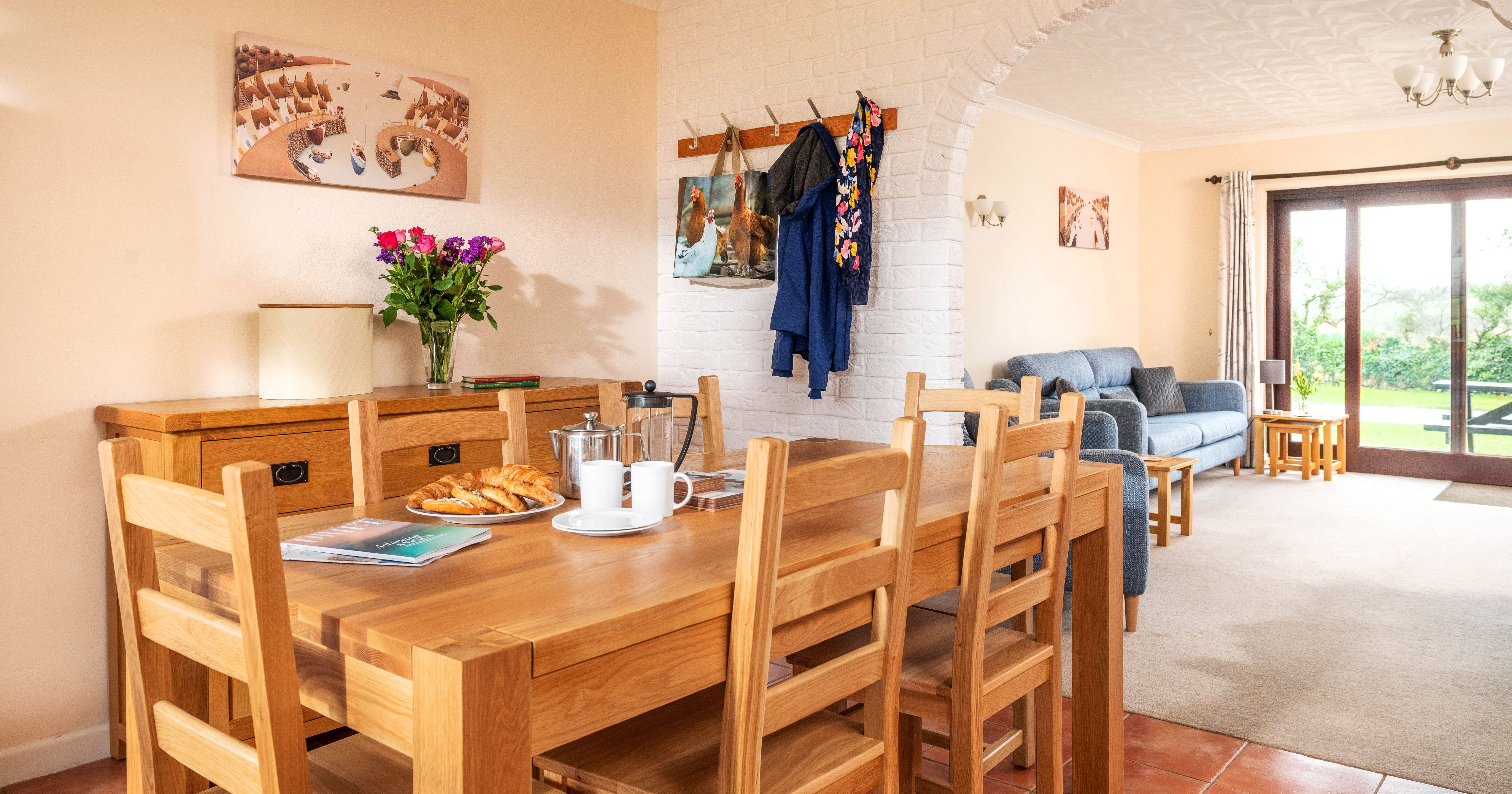 Bright open-plan dining and living room with wooden table and chairs, blue sofa, flowers on a sideboard, coat rack, and large glass doors leading to a garden.