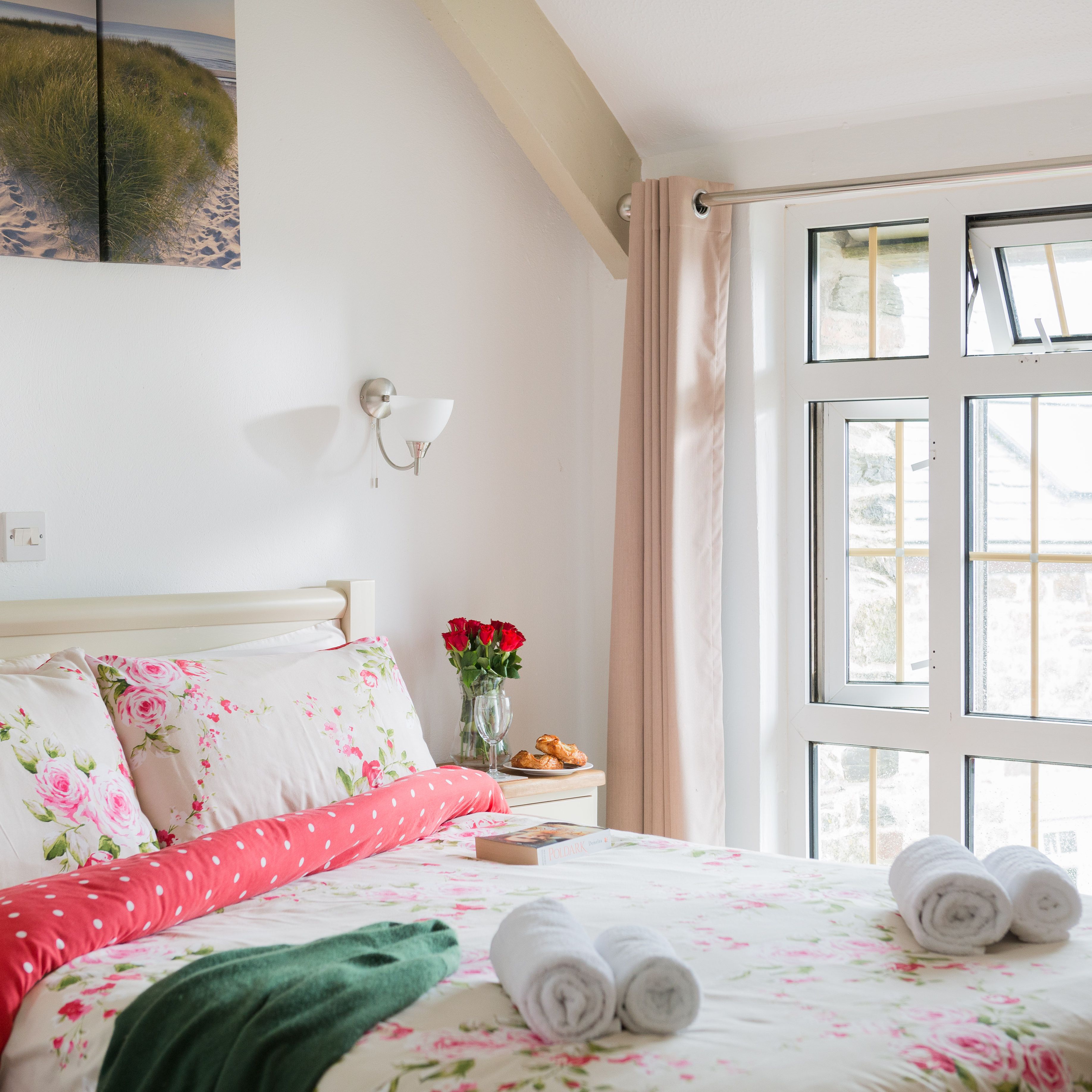 Bright and cozy bedroom with floral bedding, large window with beige curtains, bedside table with a vase of red roses, and neatly rolled towels on the bed.