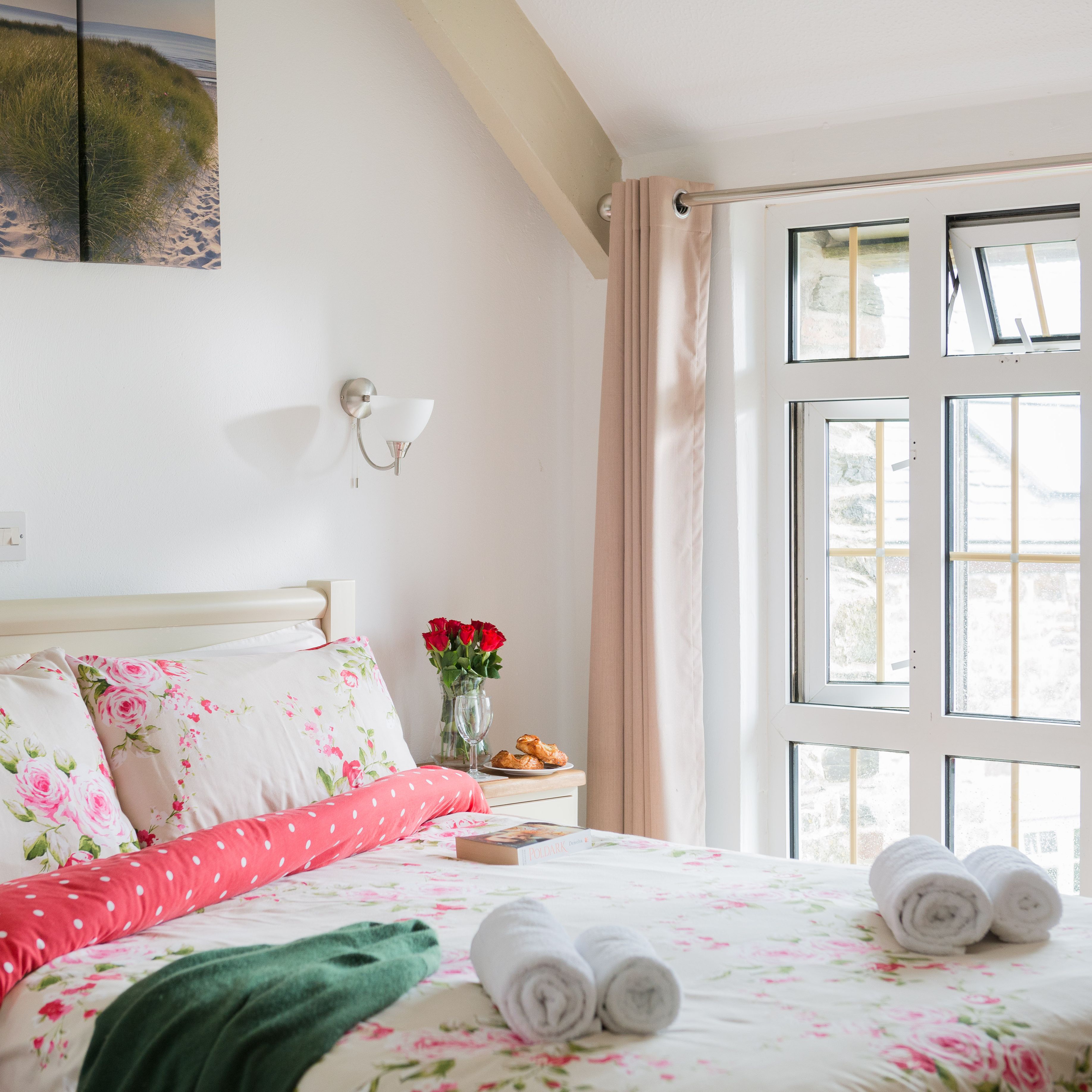 Bright and cozy bedroom with floral bedding, large window with beige curtains, bedside table with a vase of red roses, and neatly rolled towels on the bed.
