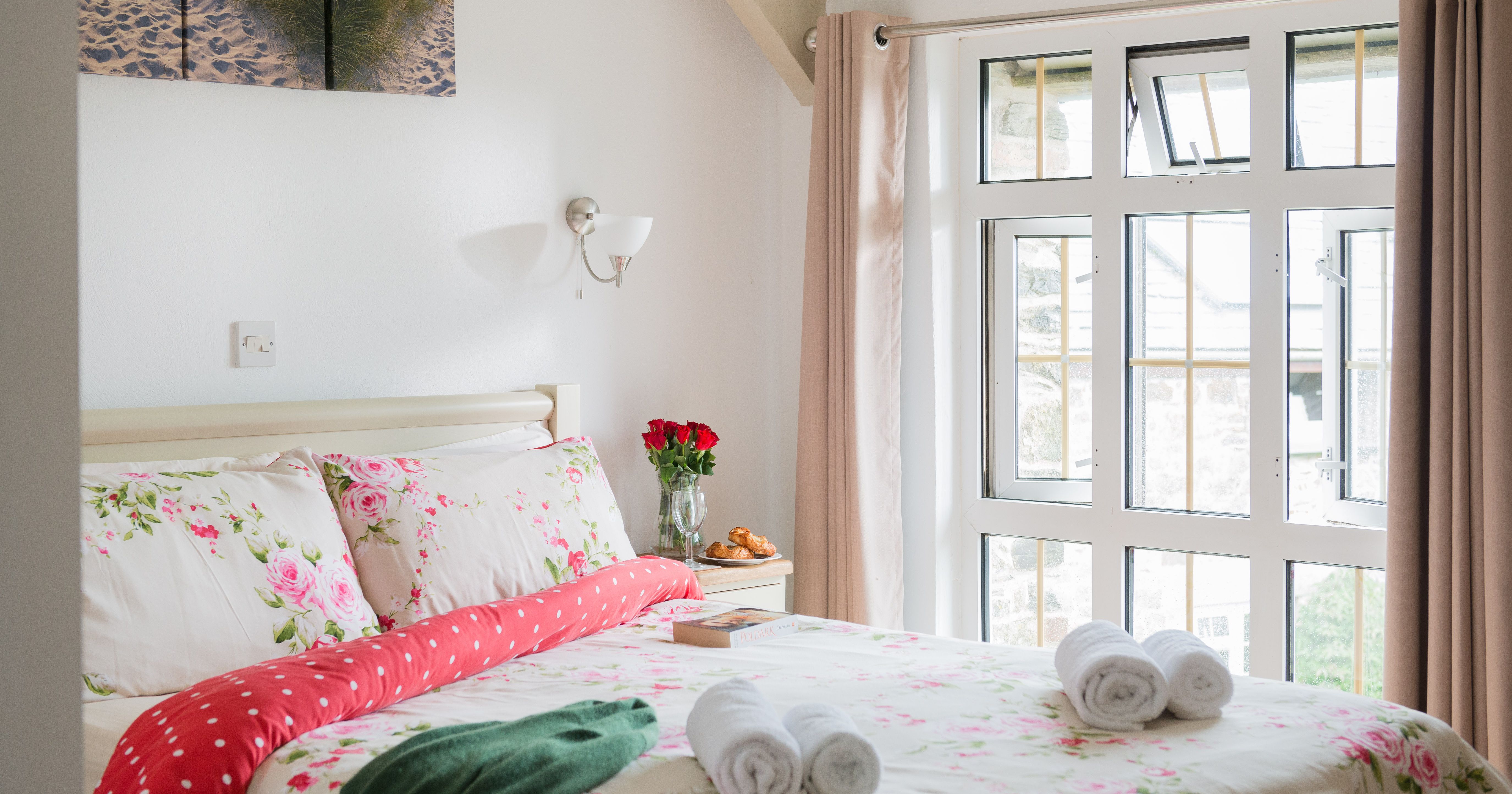Bright and cozy bedroom with floral bedding, large window with beige curtains, bedside table with a vase of red roses, and neatly rolled towels on the bed.
