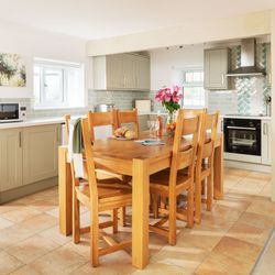Bright and modern kitchen with light green cabinets, a wooden dining table with six chairs, pink flowers in a vase, bread and fruit on the table, tiled floor, and stainless steel appliances.