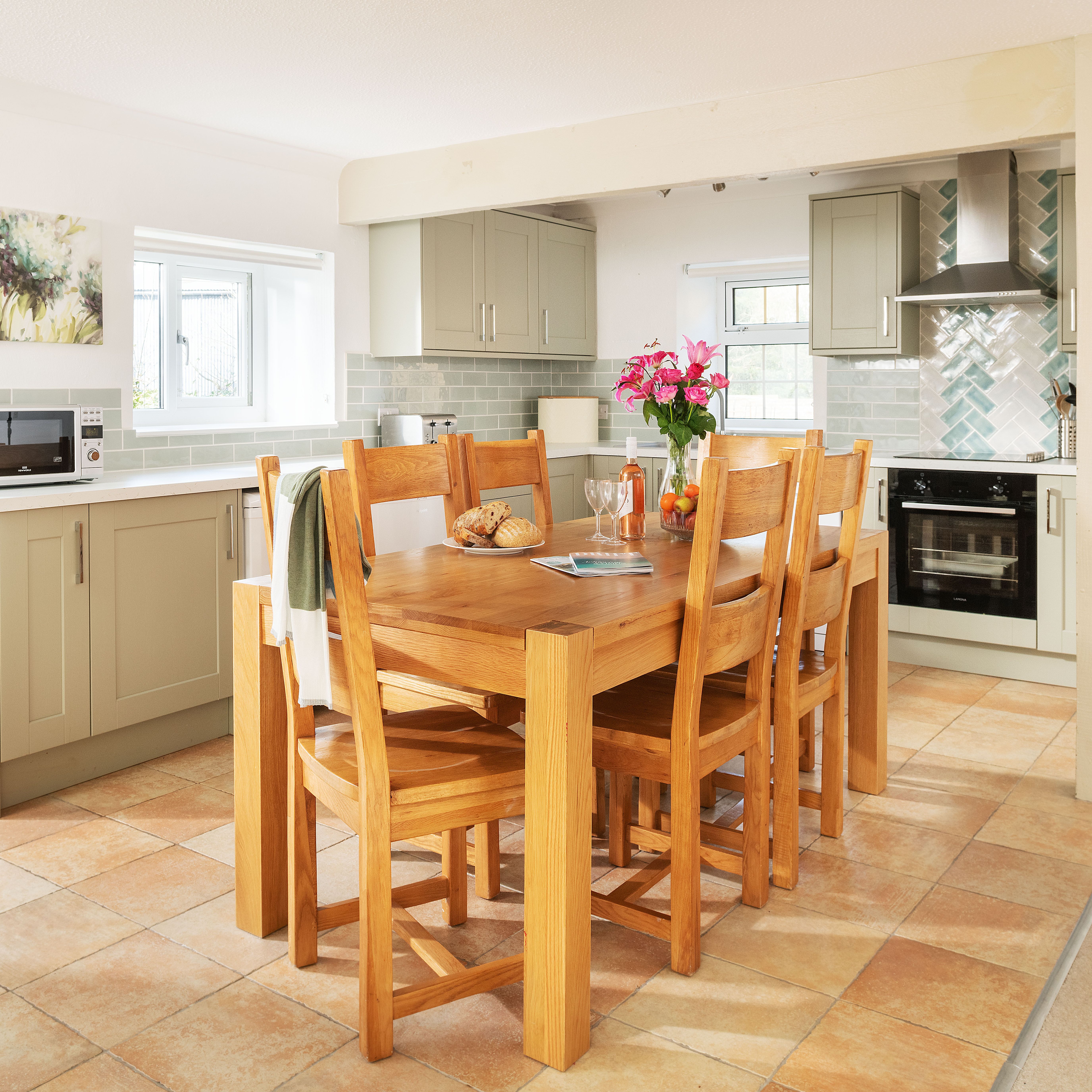 Bright and modern kitchen with light green cabinets, a wooden dining table with six chairs, pink flowers in a vase, bread and fruit on the table, tiled floor, and stainless steel appliances.