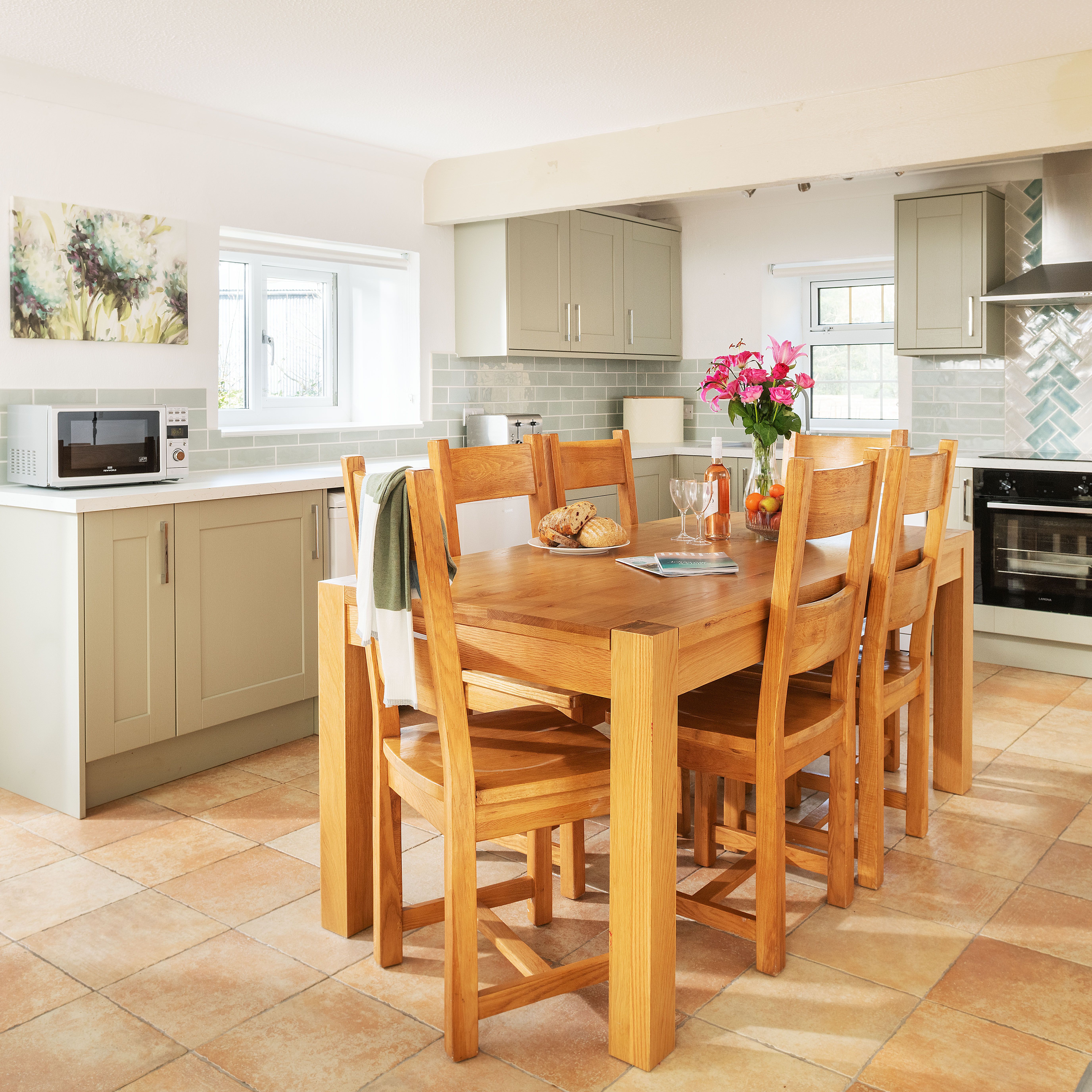 Bright and modern kitchen with light green cabinets, a wooden dining table with six chairs, pink flowers in a vase, bread and fruit on the table, tiled floor, and stainless steel appliances.