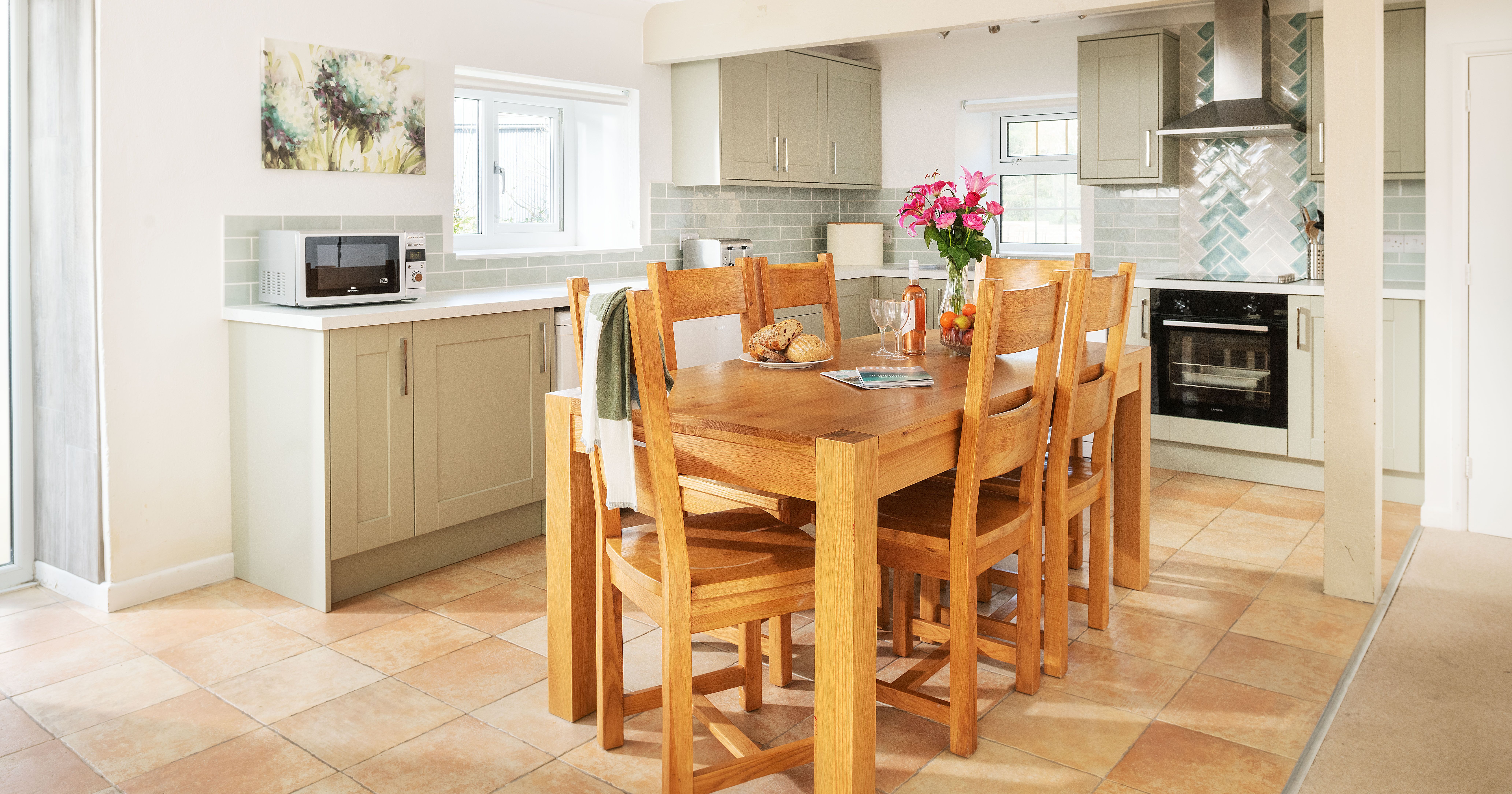 Bright and modern kitchen with light green cabinets, a wooden dining table with six chairs, pink flowers in a vase, bread and fruit on the table, tiled floor, and stainless steel appliances.