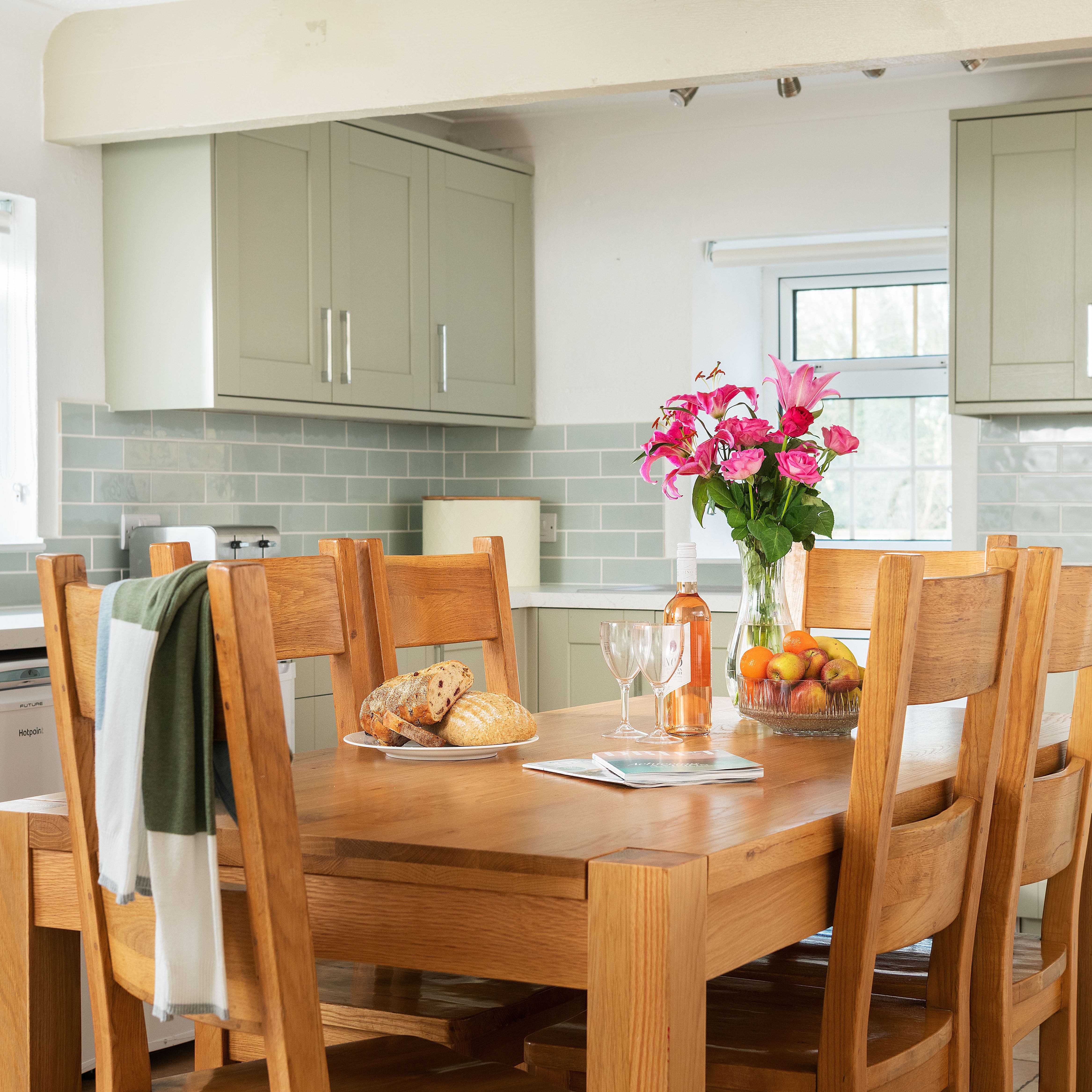 Modern kitchen with light green cabinets, wooden dining table set with bread, fruit, flowers, and a bottle of rosé wine. Natural light coming through windows and tiled backsplash visible.
