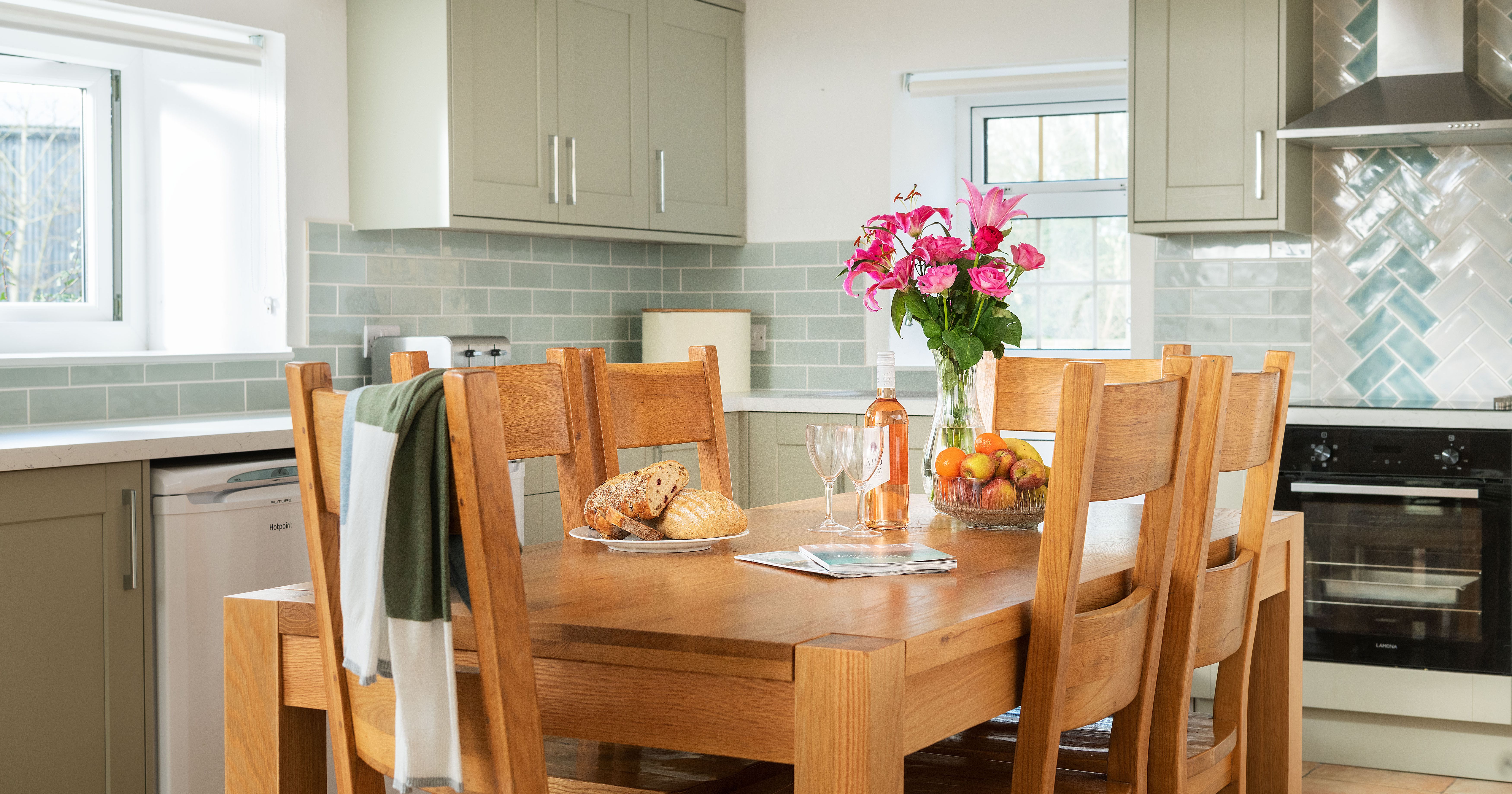 Modern kitchen with light green cabinets, wooden dining table set with bread, fruit, flowers, and a bottle of rosé wine. Natural light coming through windows and tiled backsplash visible.