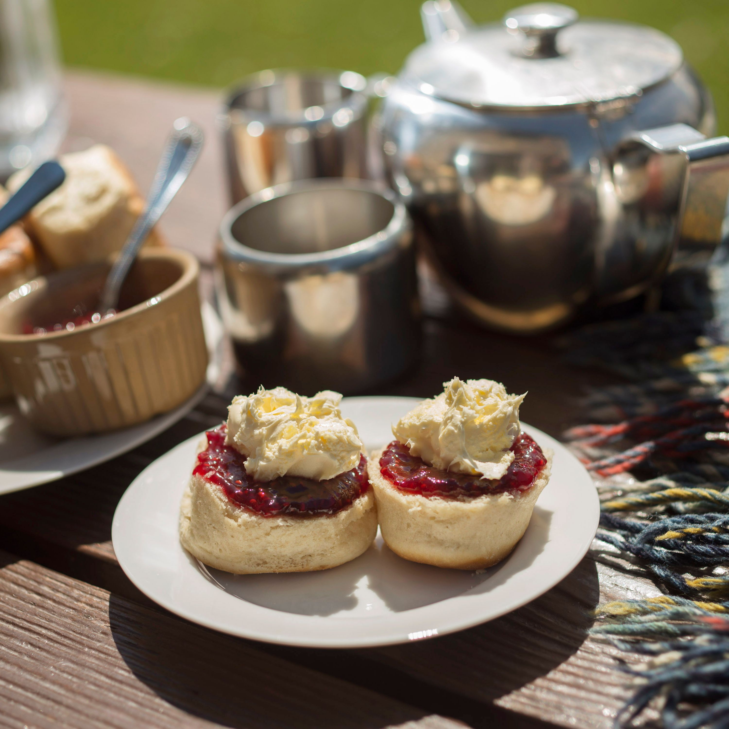 Outdoor afternoon tea setup with scones topped with clotted cream and jam, a silver teapot, cups, and a colorful plaid blanket on a wooden table.