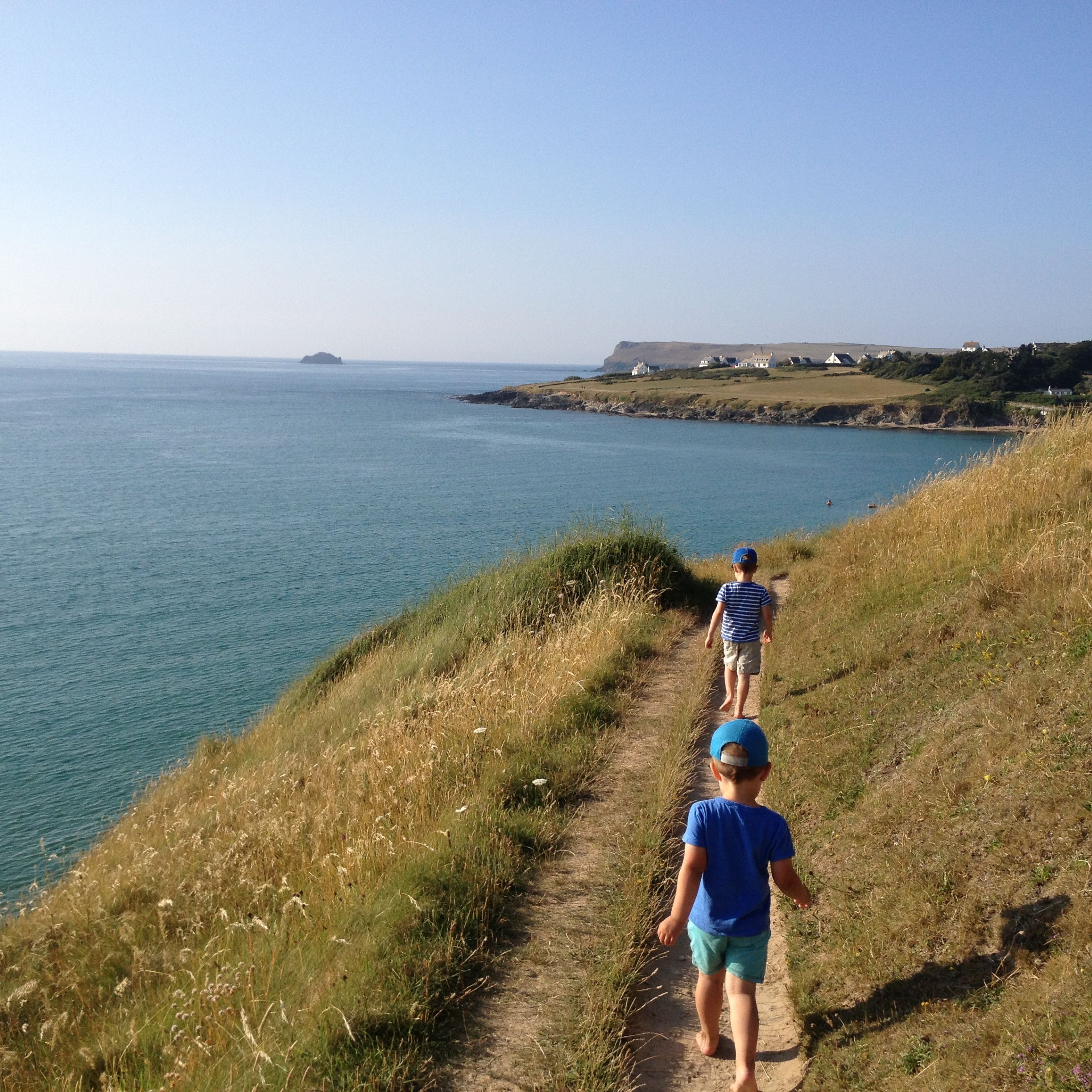 Two children walking barefoot on a narrow dirt path along a grassy coastal cliff overlooking the sea, with distant houses and clear blue sky.