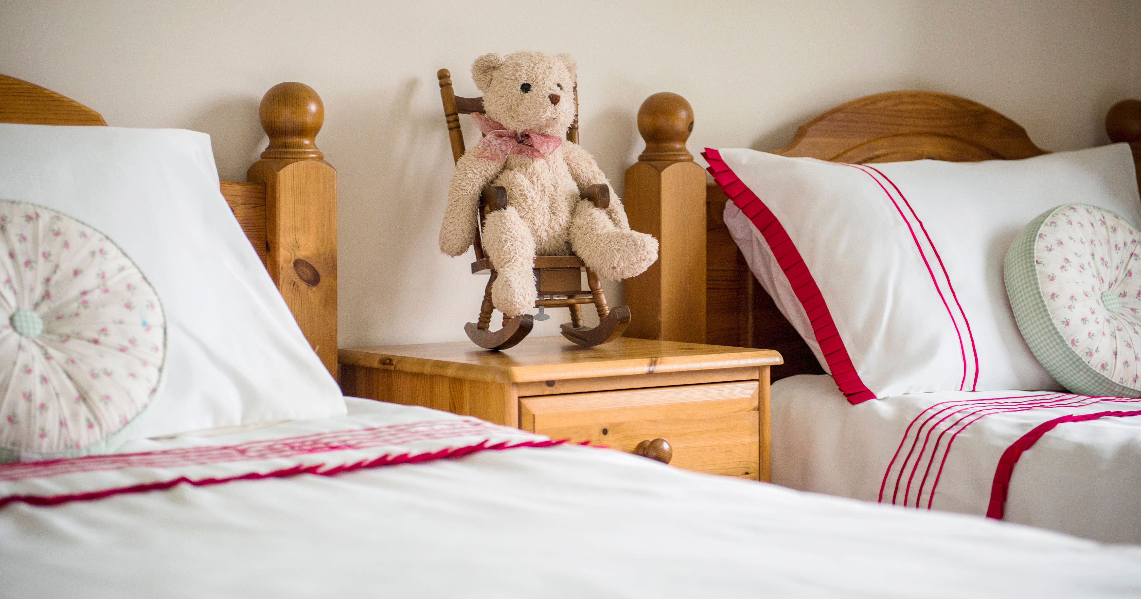 A cozy bedroom with two single beds, each with white bedding and red accents, and a wooden nightstand in the middle holding a teddy bear sitting on a small wooden rocking chair.