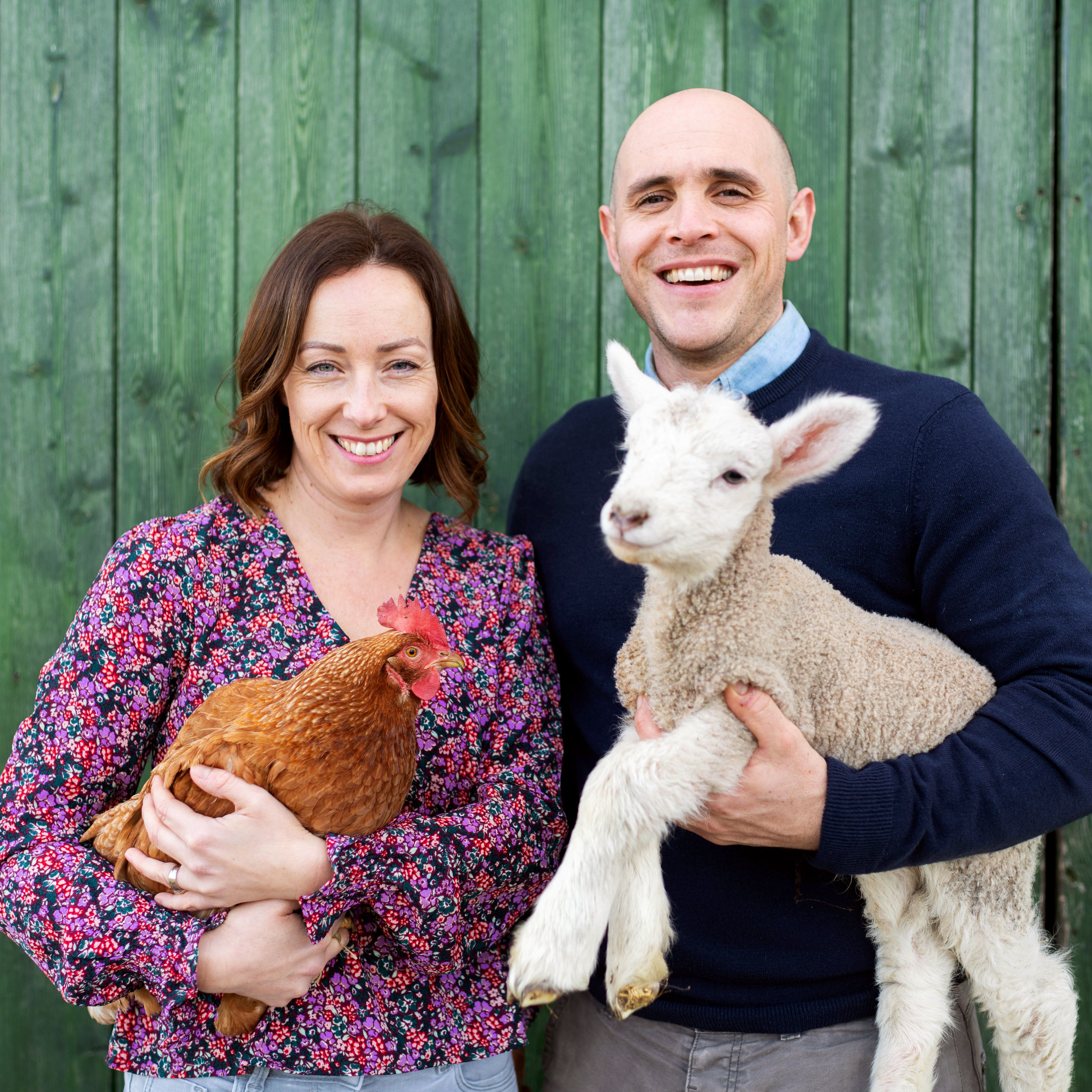 Smiling man and woman standing in front of a green wooden wall. The woman is holding a brown chicken and the man is holding a lamb.