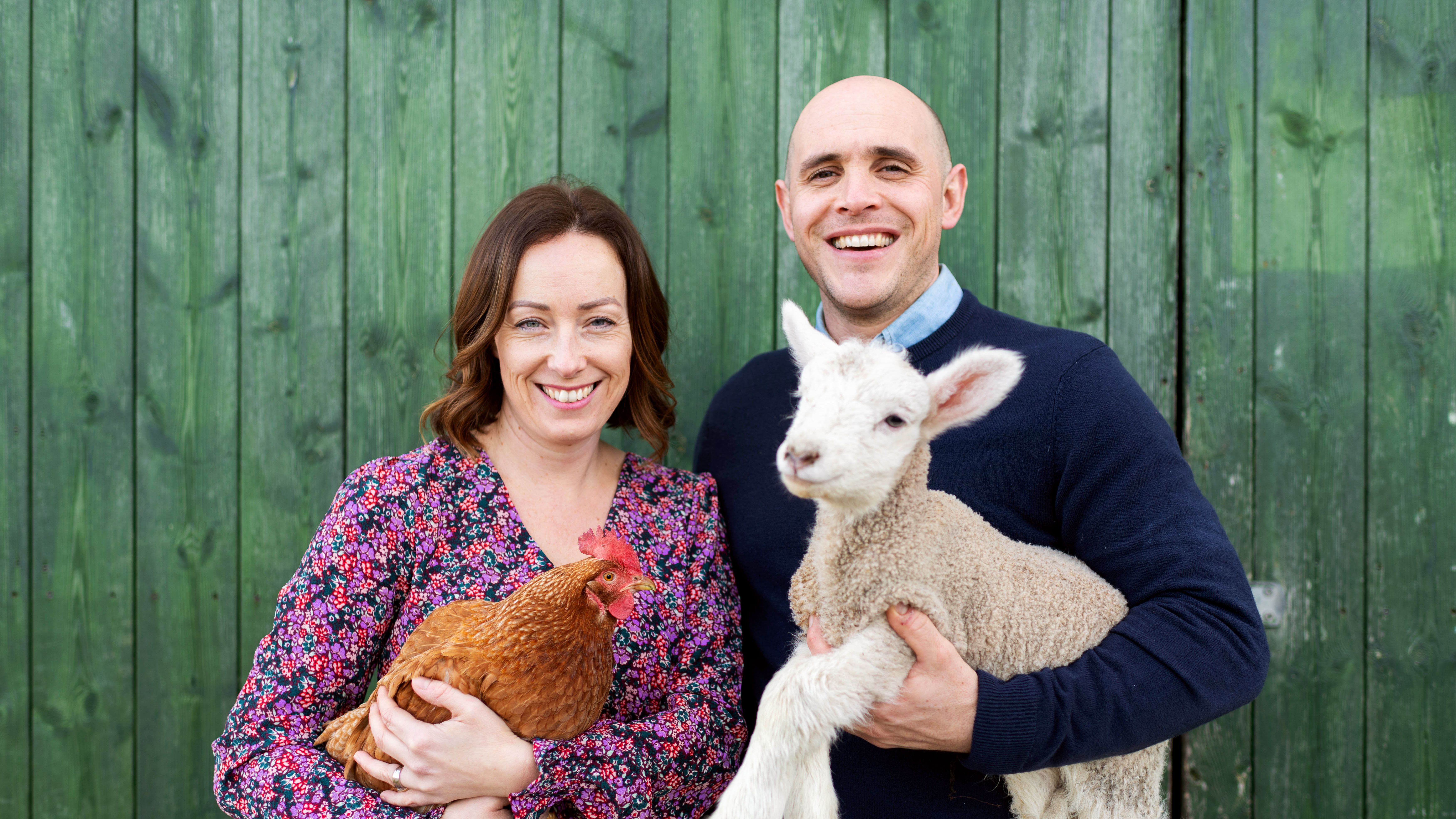 Smiling man and woman standing in front of a green wooden wall. The woman is holding a brown chicken and the man is holding a lamb.