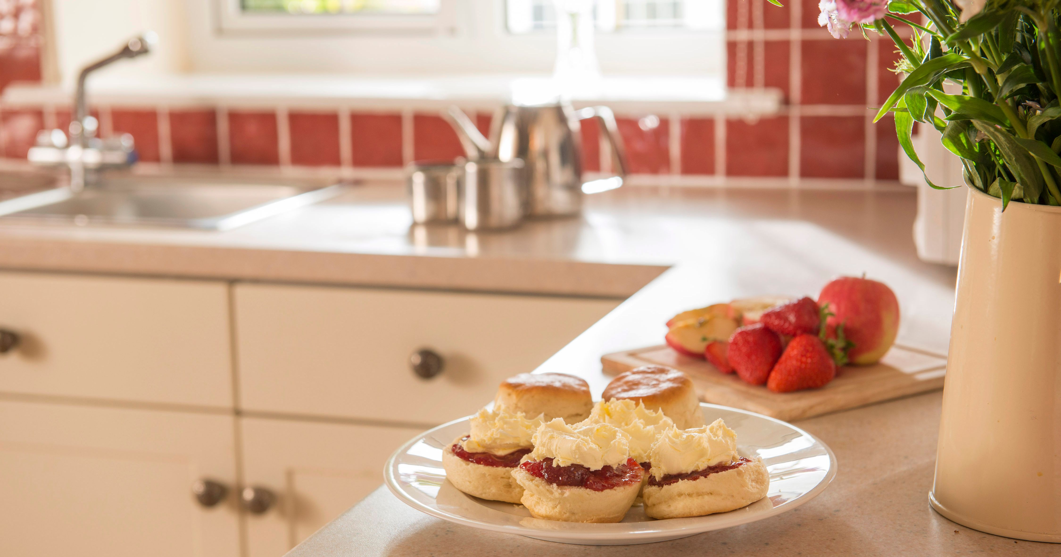 A plate of scones with jam and clotted cream on a kitchen counter, with a vase of flowers, strawberries, and an apple in the background.