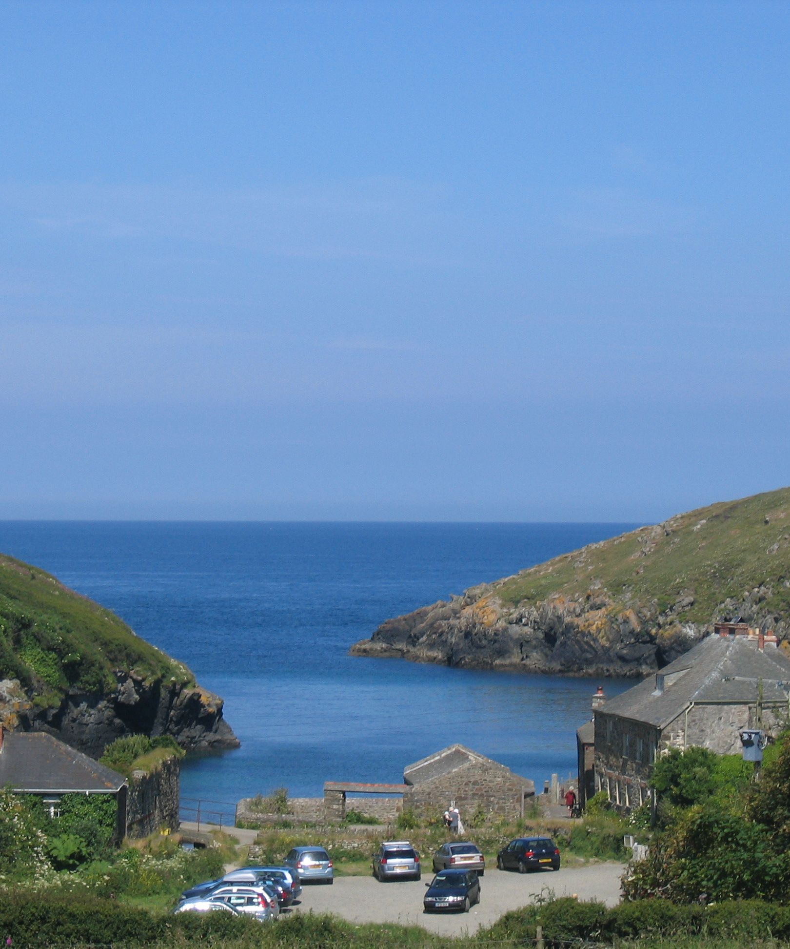A scenic coastal village with stone houses, a small parking area with cars, green hills on both sides, and a calm blue sea in the background under a clear sky.