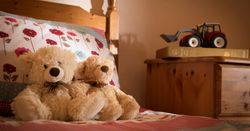 Two teddy bears sitting on a bed with floral bedding, next to a wooden nightstand with a toy tractor on top of a book.