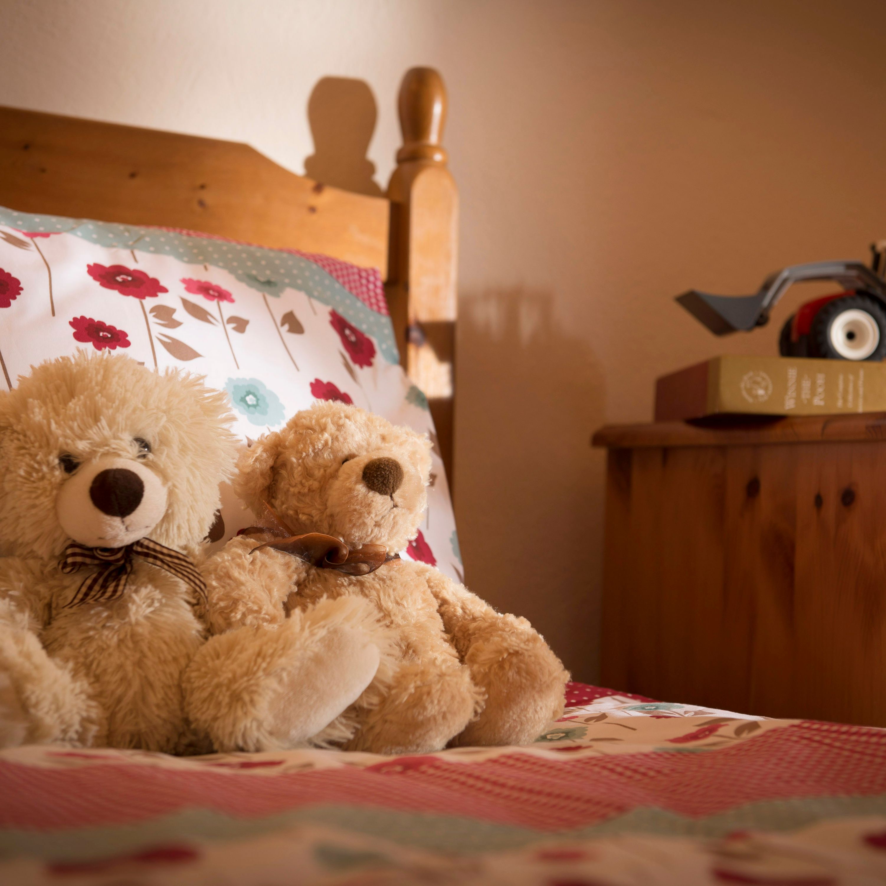 Two teddy bears sitting on a bed with floral bedding, next to a wooden nightstand with a toy tractor on top of a book.