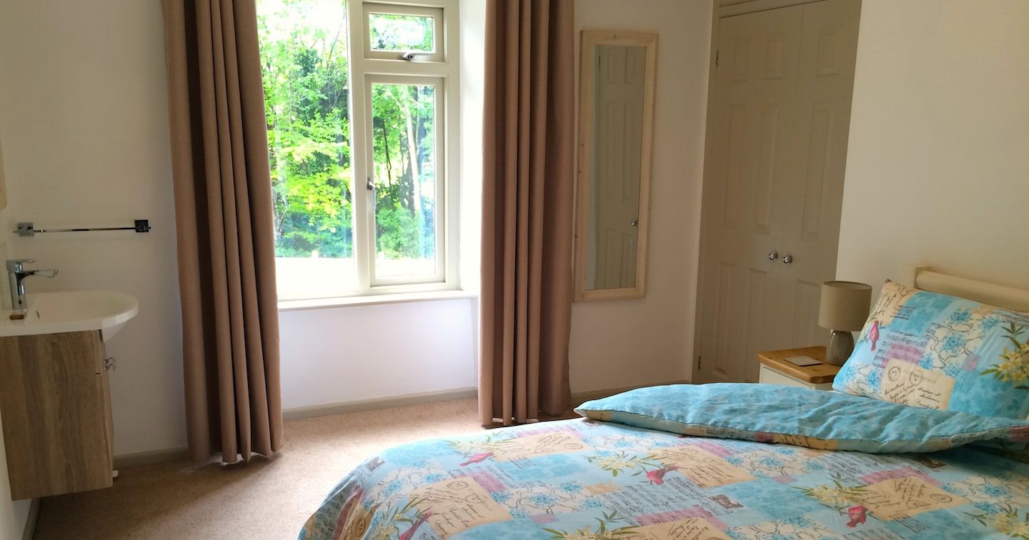 A cozy bedroom with a large window, beige curtains, a bed covered with a colorful duvet, a bedside table with a lamp, a wall mirror, built-in wardrobes, and a small wall-mounted sink.