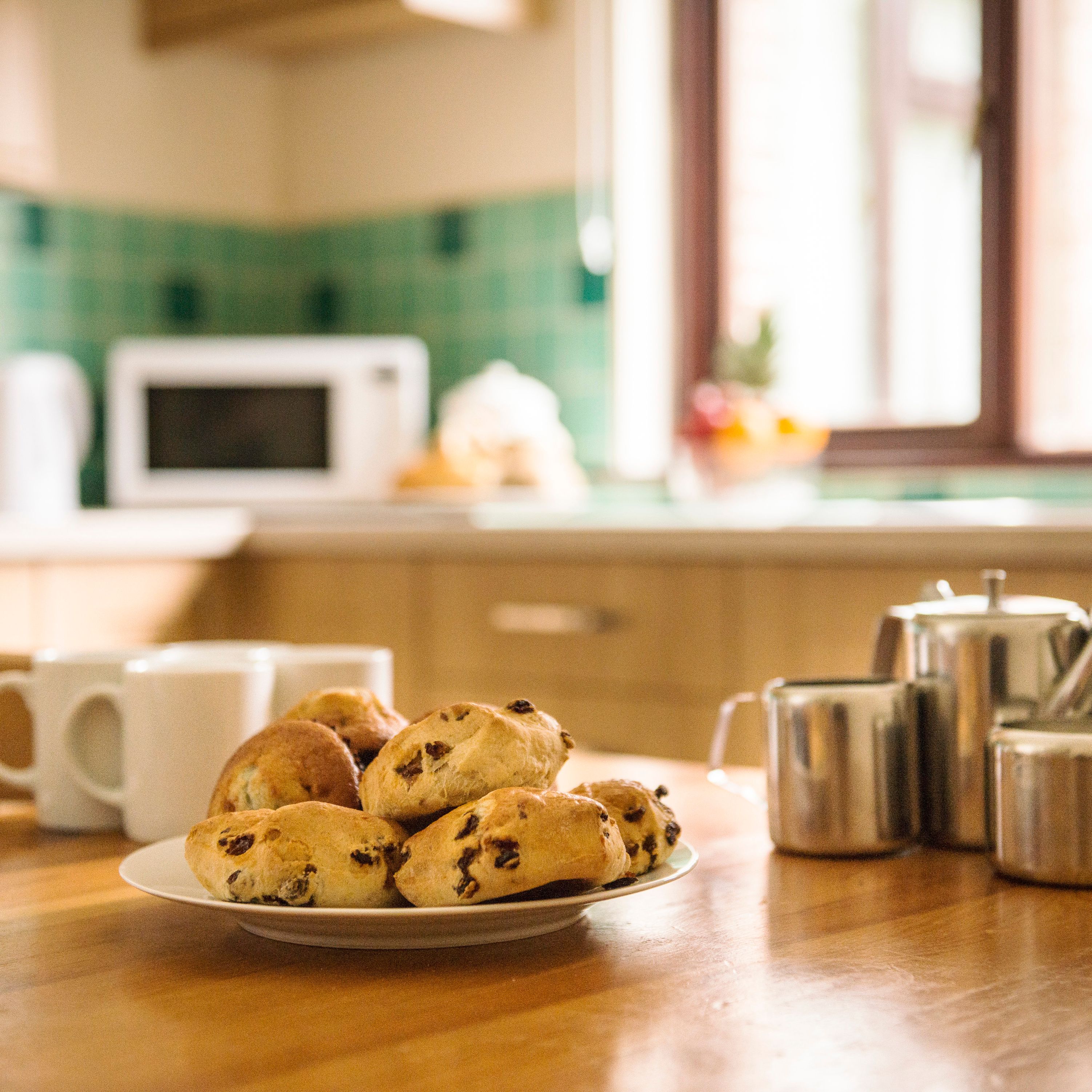 A plate of scones on a wooden kitchen table with three white mugs and a stainless steel tea set, in a cozy kitchen with a microwave and green-tiled backsplash in the background.