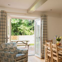 Bright dining room with wooden table and chairs, an armchair with a geometric pattern, open patio doors with plaid curtains, and an outdoor picnic table in a sunny garden.