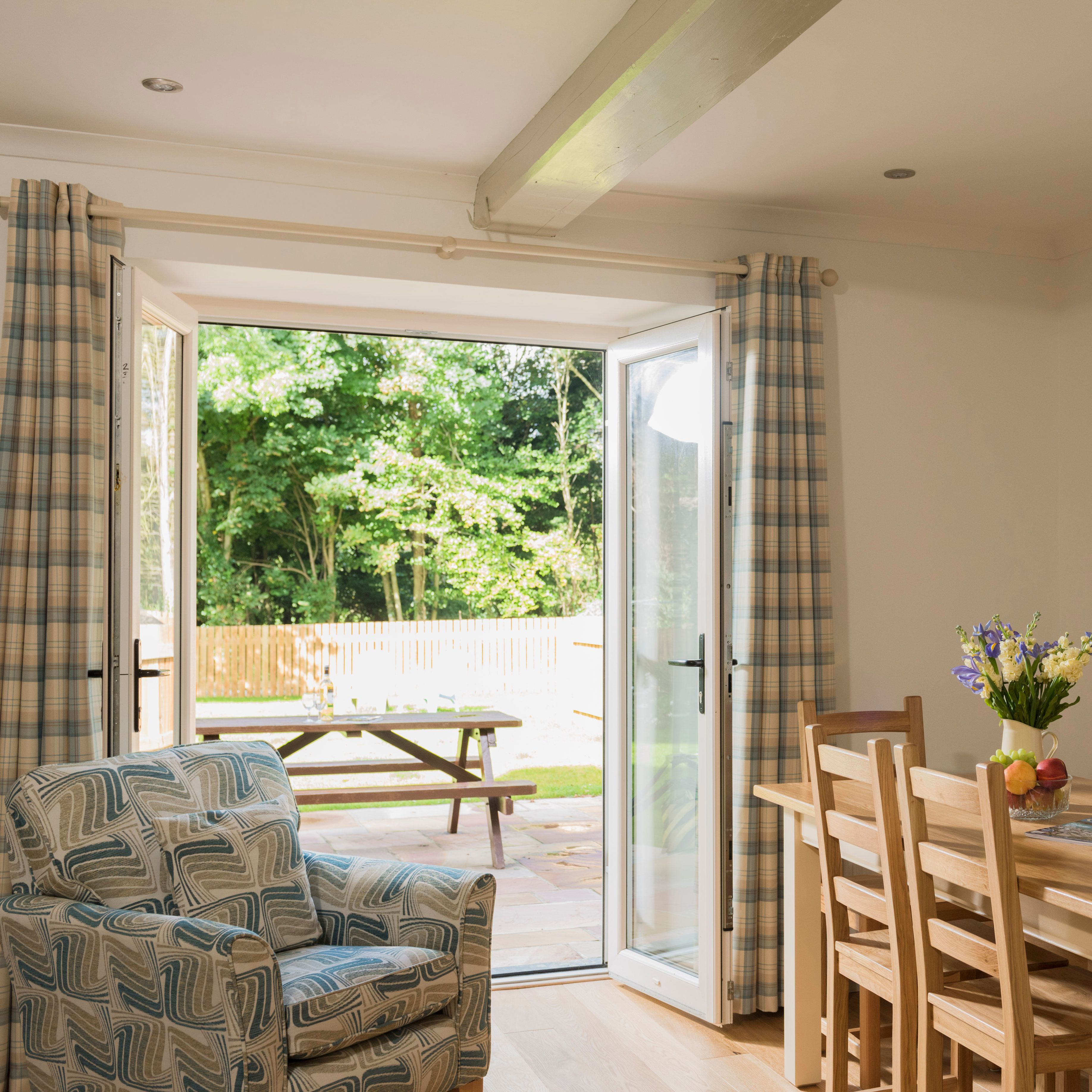 Bright dining room with wooden table and chairs, an armchair with a geometric pattern, open patio doors with plaid curtains, and an outdoor picnic table in a sunny garden.