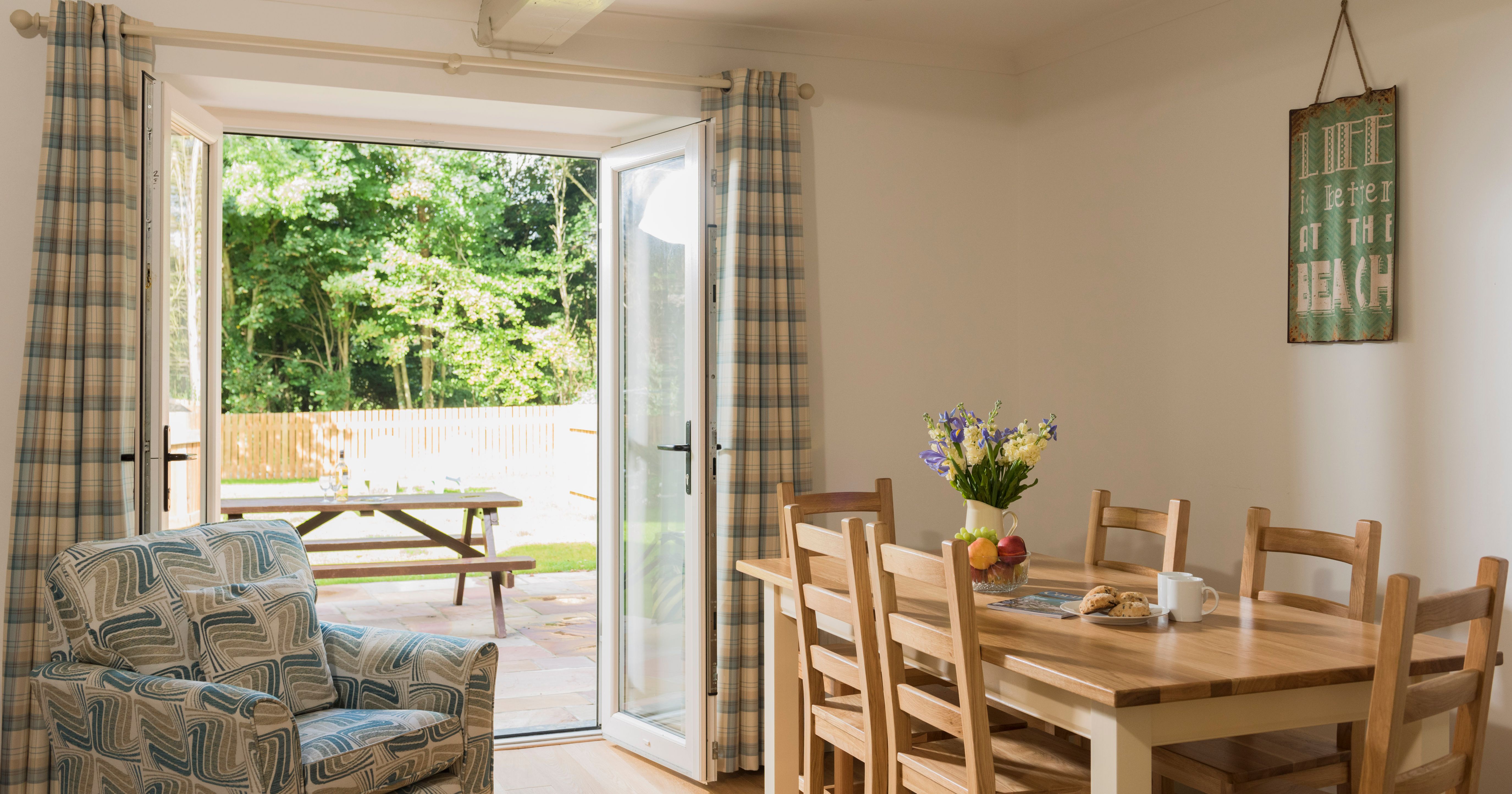 Bright dining room with wooden table and chairs, an armchair with a geometric pattern, open patio doors with plaid curtains, and an outdoor picnic table in a sunny garden.