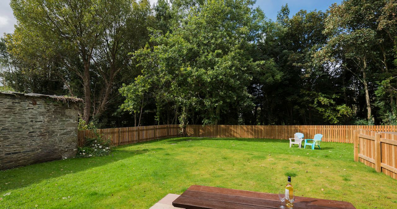 A fenced backyard with green grass, a wooden picnic table with a bottle and two glasses on it, three chairs in the distance, and trees surrounding the yard.