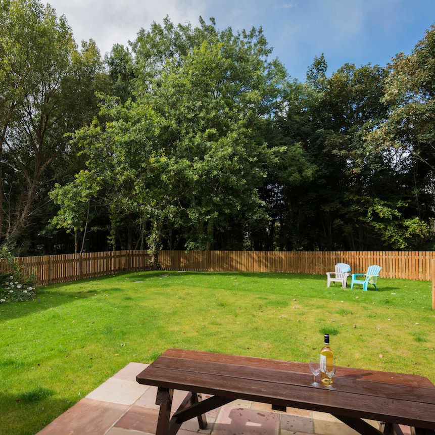 A fenced backyard with green grass, a wooden picnic table with a bottle and two glasses on it, three chairs in the distance, and trees surrounding the yard.