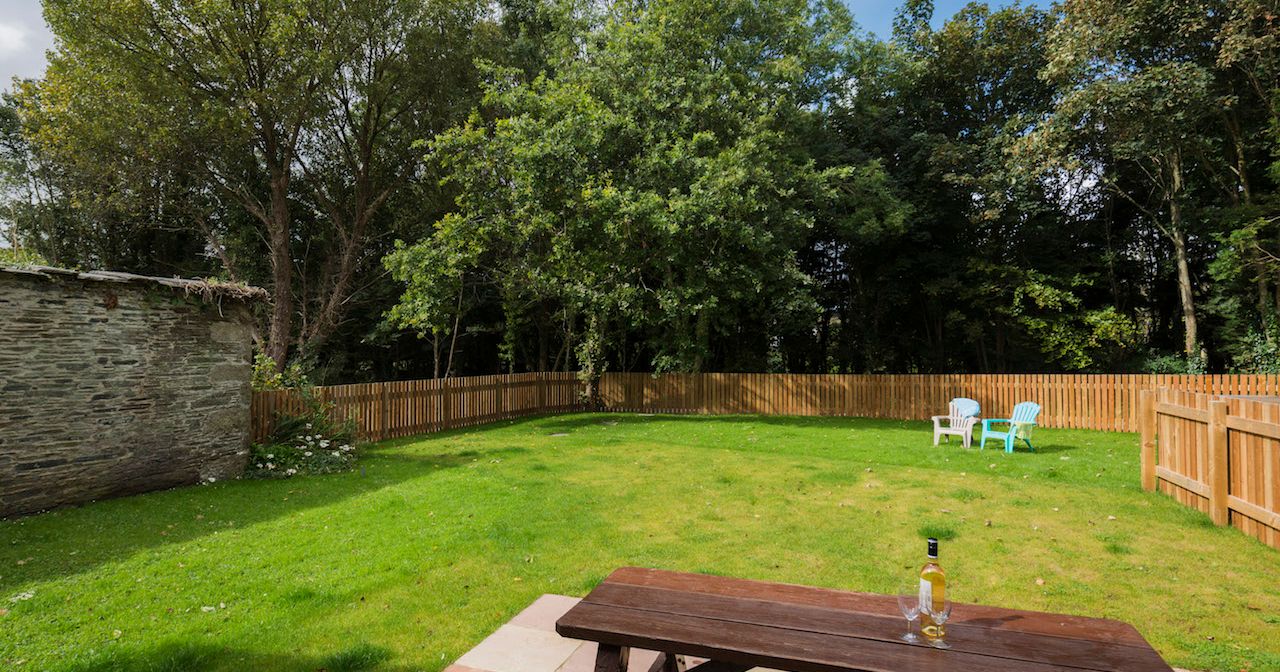 A fenced backyard with green grass, a wooden picnic table with a bottle and two glasses on it, three chairs in the distance, and trees surrounding the yard.