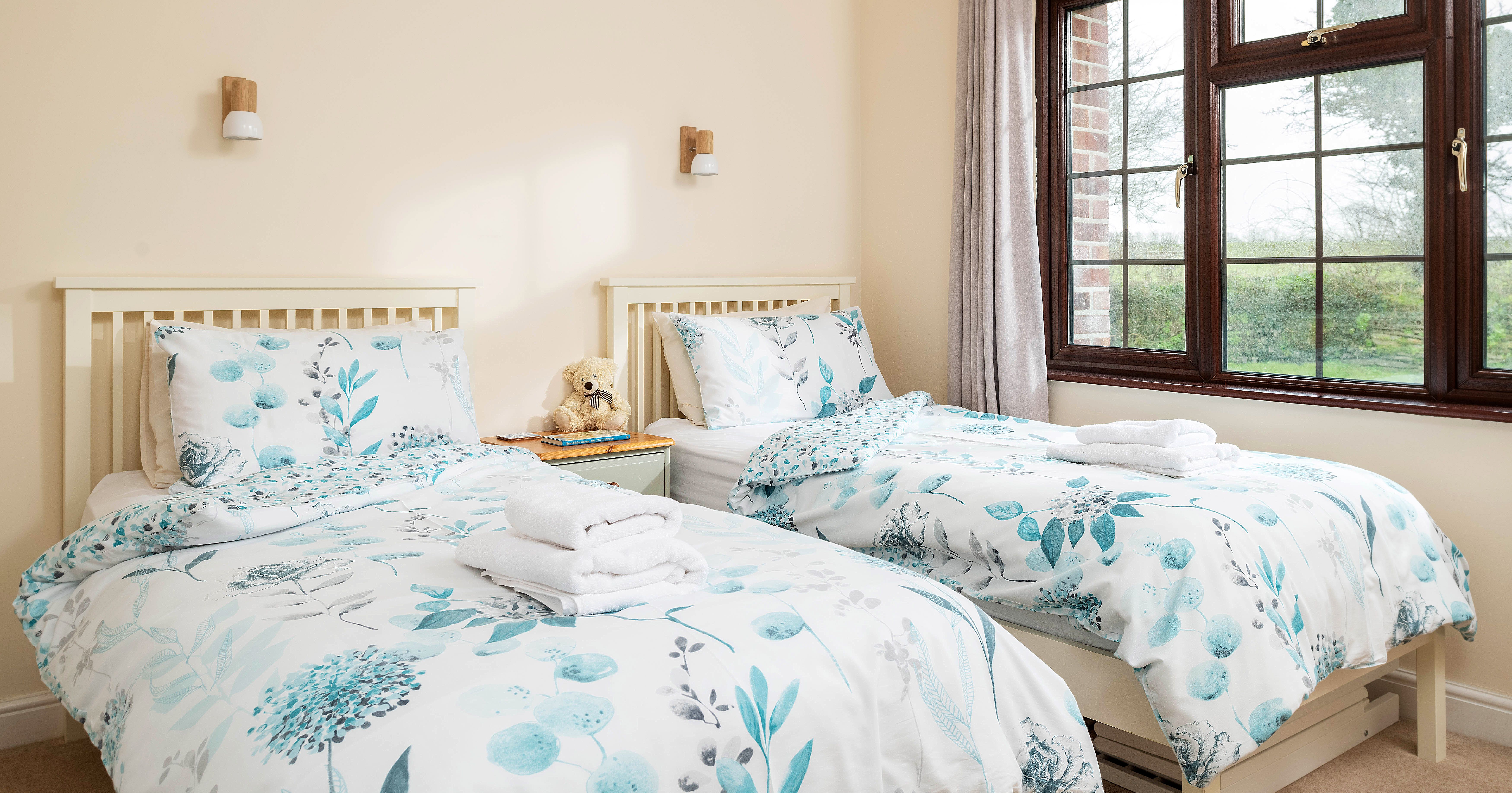 Bright bedroom with two single beds featuring white and blue floral bedding, a bedside table with a teddy bear and books, and a large window with garden view.