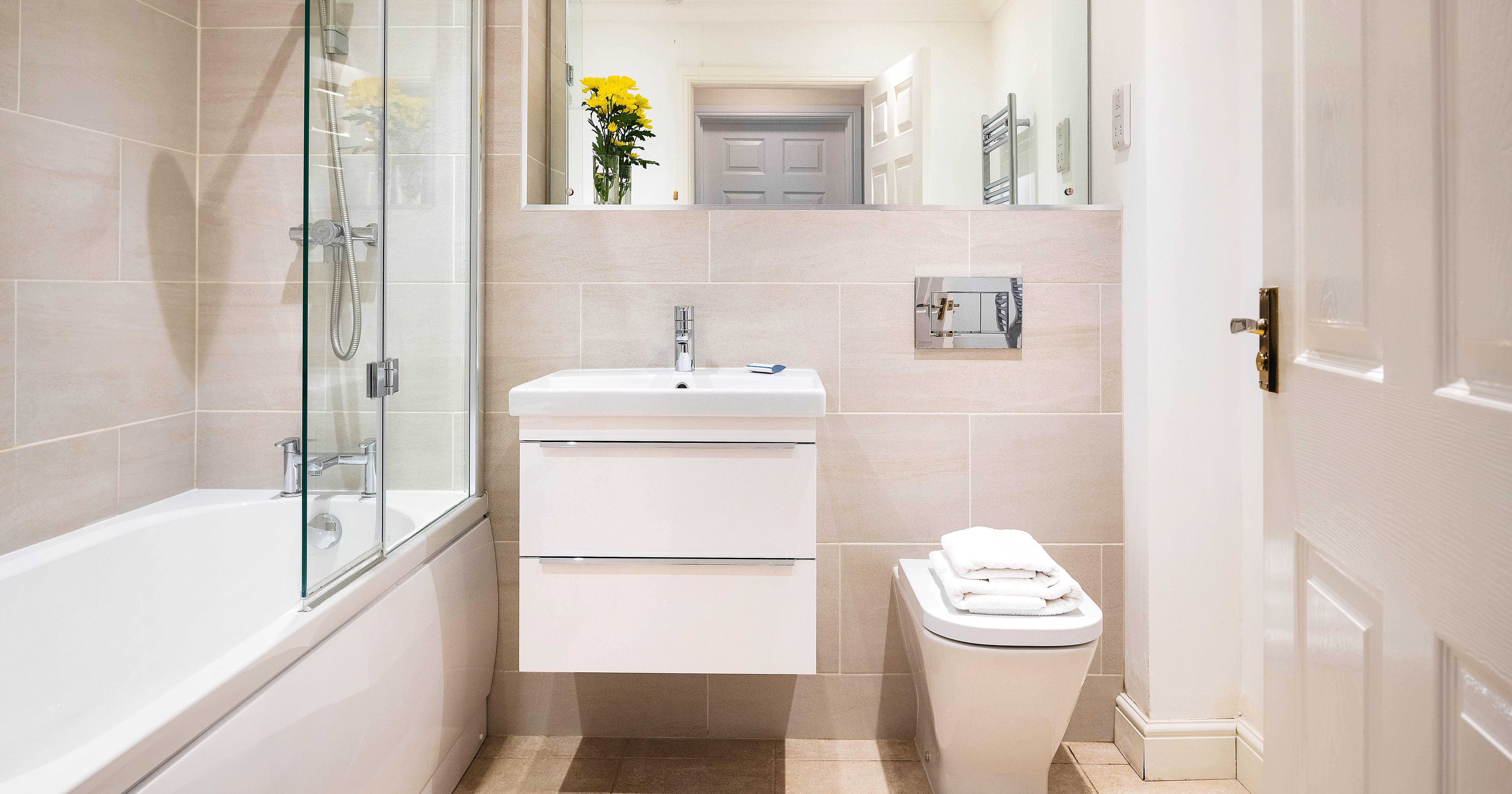Modern bathroom with beige tiles, a glass shower over a bathtub, a floating vanity with sink, a bidet with folded towels, large mirror, and a vase with yellow flowers.