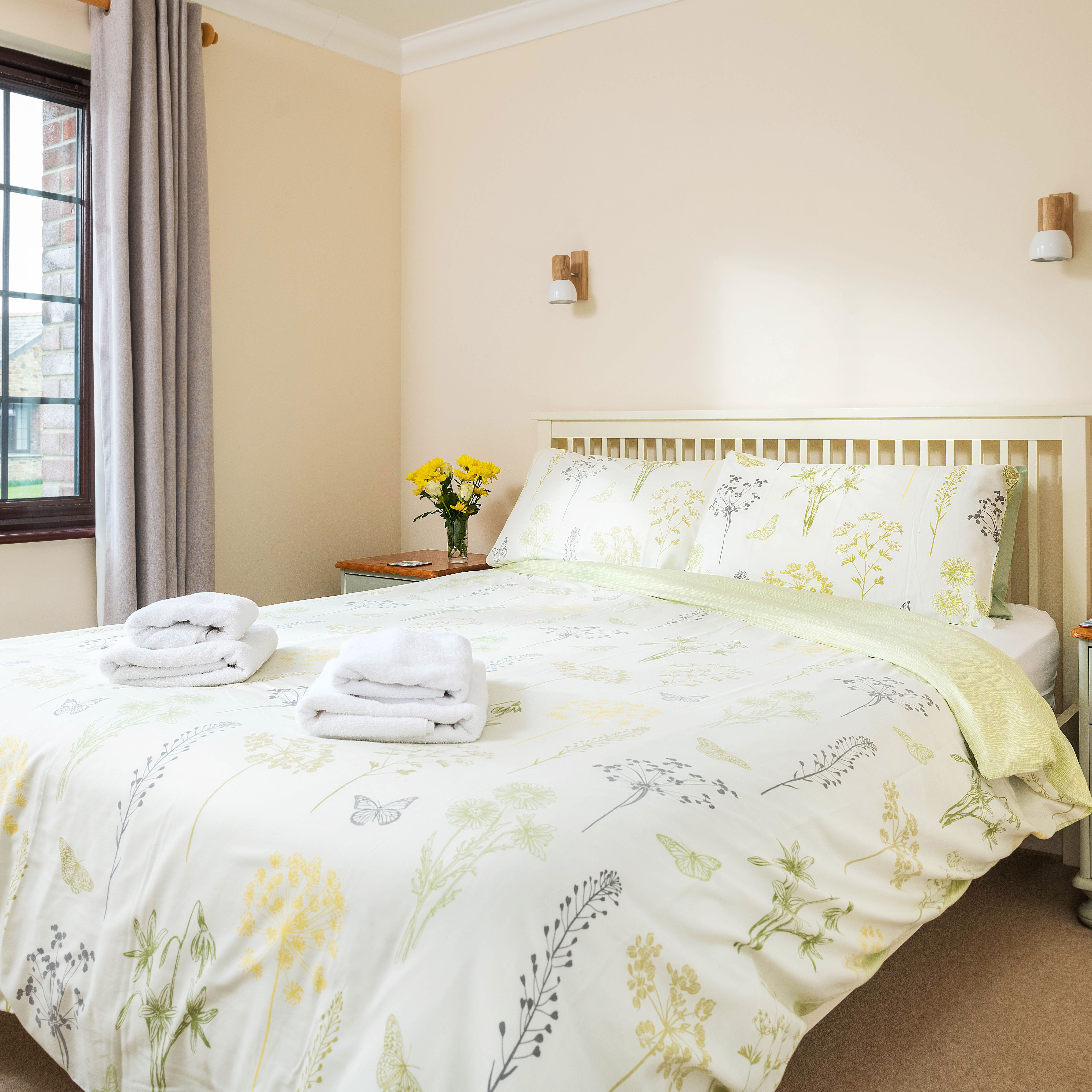 Bright and cozy bedroom with floral bedding, neatly folded towels on the bed, two bedside tables with flowers, books, and a mug, and a large window letting in natural light.
