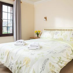 Bright and cozy bedroom with floral bedding, neatly folded towels on the bed, two bedside tables with flowers, books, and a mug, and a large window letting in natural light.