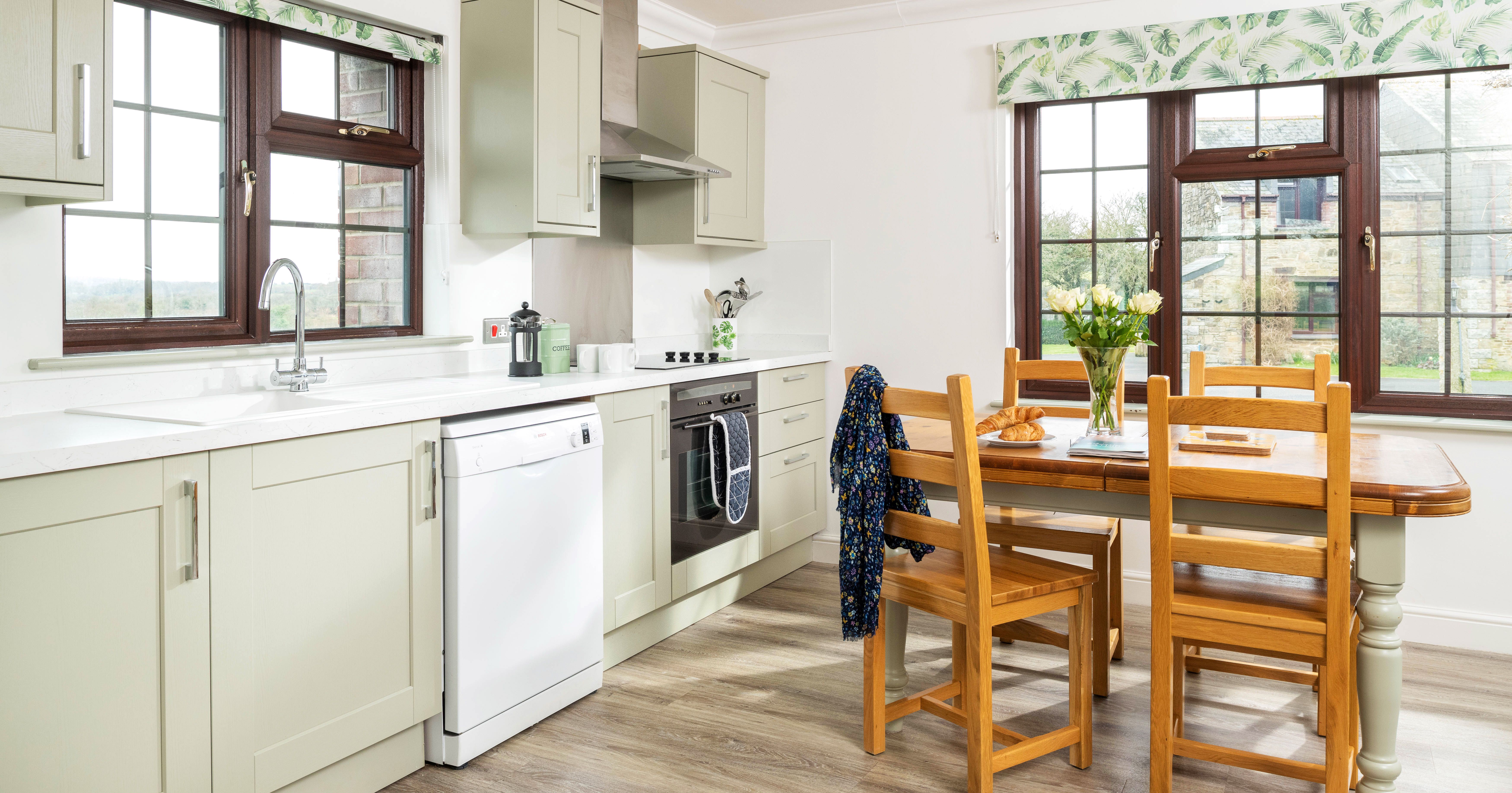 Bright and modern kitchen interior with light green cabinets, wooden dining table with chairs, large windows, and a vase of flowers on the table.