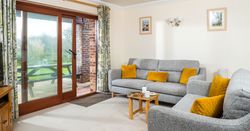 Cozy living room with grey sofas, yellow cushions, wooden coffee table, sliding glass doors, floral curtains, and a view of a garden with a picnic table outside.