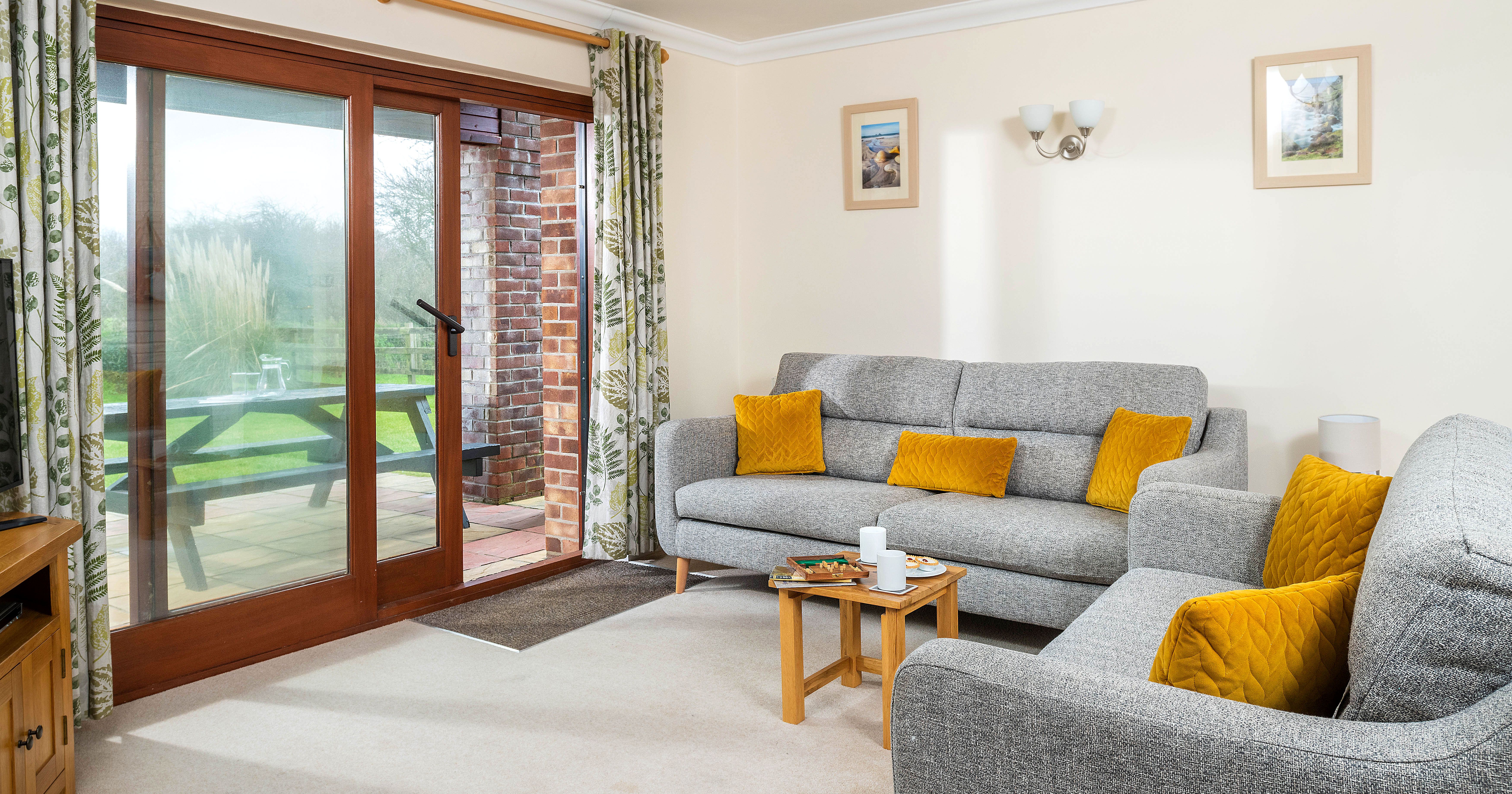 Cozy living room with grey sofas, yellow cushions, wooden coffee table, sliding glass doors, floral curtains, and a view of a garden with a picnic table outside.