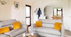 Bright living room with two gray sofas featuring yellow cushions, a small wooden coffee table with mugs and snacks, and a view into the kitchen and dining area through a white brick archway.