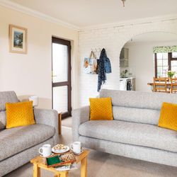 Bright living room with two gray sofas featuring yellow cushions, a small wooden coffee table with mugs and snacks, and a view into the kitchen and dining area through a white brick archway.