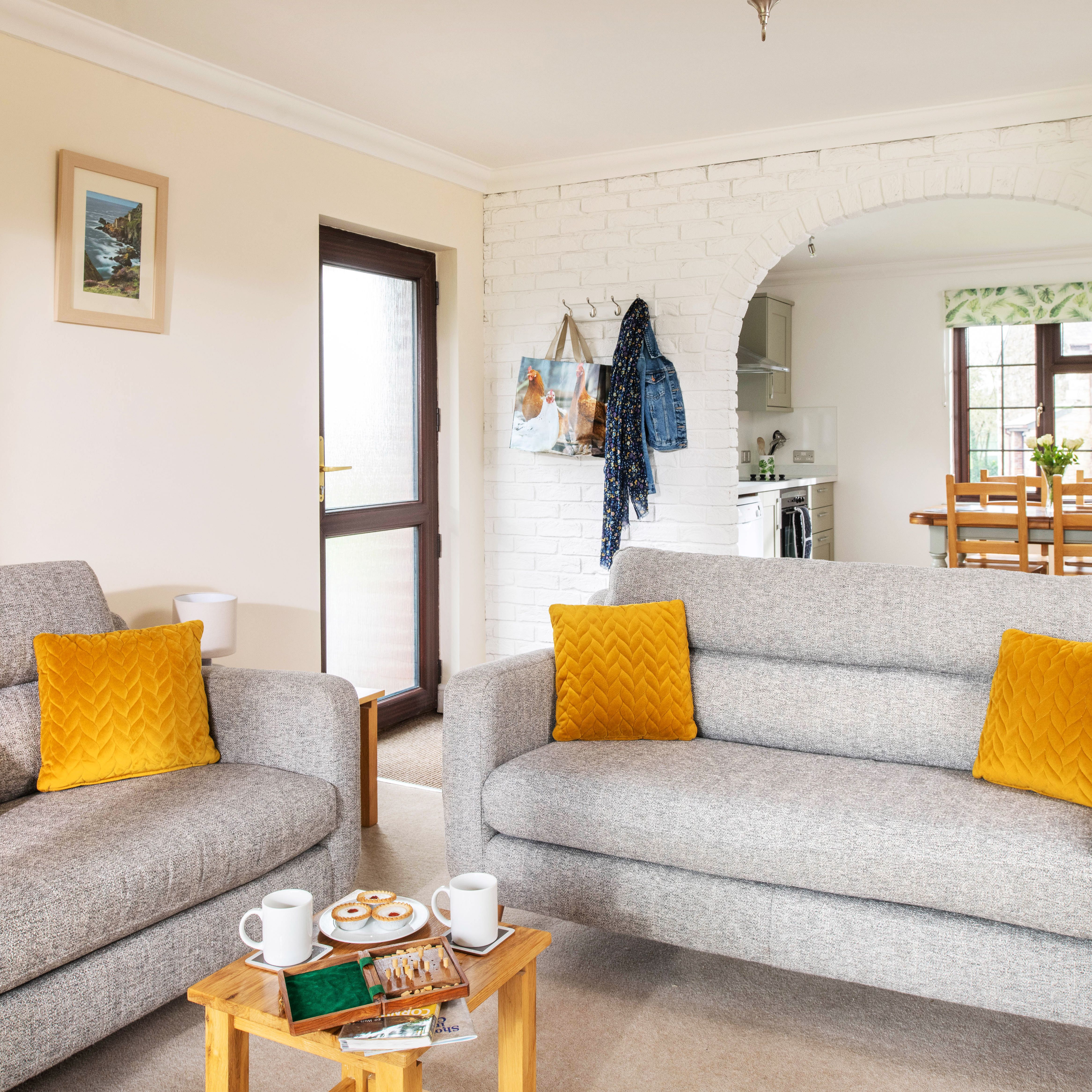 Bright living room with two gray sofas featuring yellow cushions, a small wooden coffee table with mugs and snacks, and a view into the kitchen and dining area through a white brick archway.