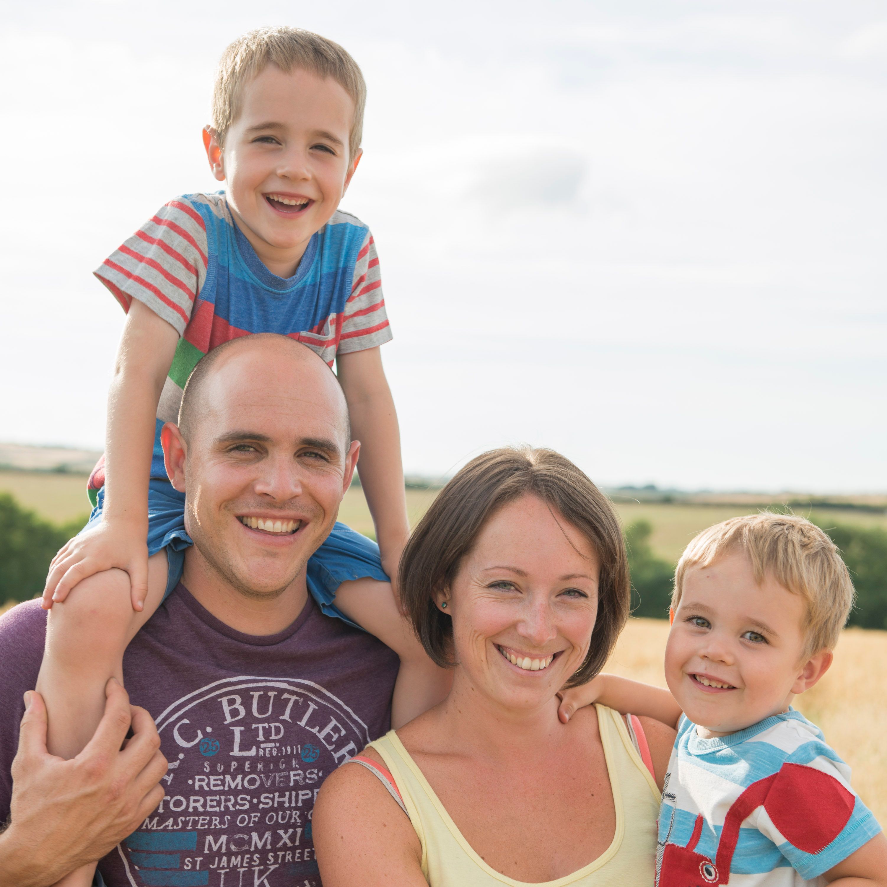 A happy family of four posing outdoors in a sunny field. The father is carrying an older boy on his shoulders, and the mother is holding a younger boy. Everyone is smiling at the camera.