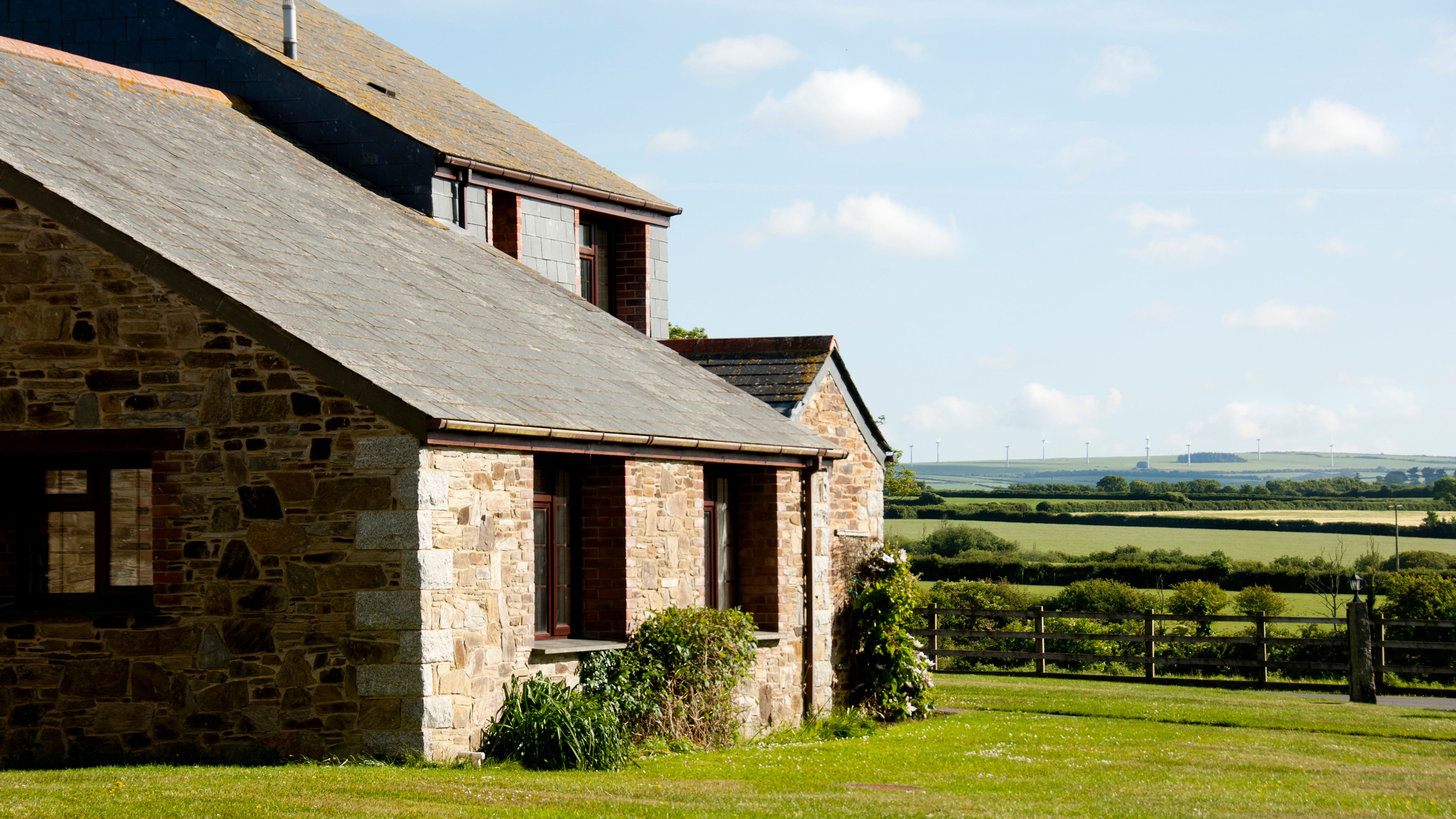 A stone farmhouse with a slate roof in a rural countryside setting, surrounded by green fields under a partly cloudy blue sky.